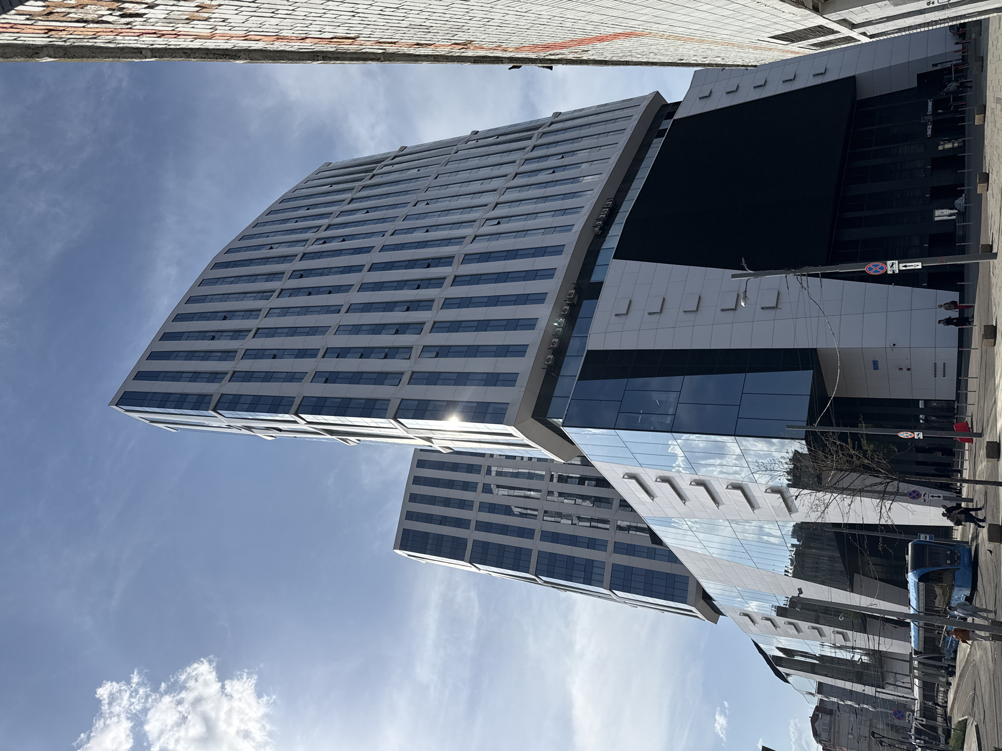 The image presents a view of a modern, multi-story building with a curved facade and glass windows, likely located in an urban environment. The structure is composed of white and dark elements. The top left corner of the frame shows a small portion of a brick wall. The sky above is blue with some clouds. A street scene is visible, with pedestrians, a blue transportation vehicle, and street signs, possibly indicating a road or public transit area. The image is taken at an unusual angle, tilted from the horizontal plane. The location is in the city of Moscow, Russia.