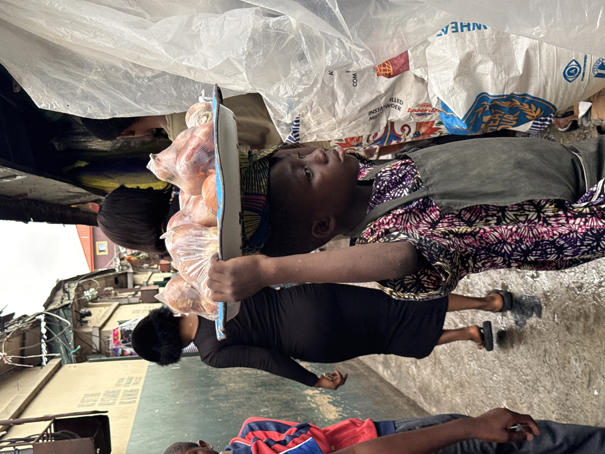 The image shows a young child carrying a tray of onions in a crowded street in Lekki, Nigeria. The child is wearing a patterned shirt and a head wrap. An adult woman in a black dress walks nearby. The surroundings include a mix of buildings, bags of wheat, and other people. The overall scene indicates a bustling marketplace environment. The child is holding the tray of onions aloft and steady.