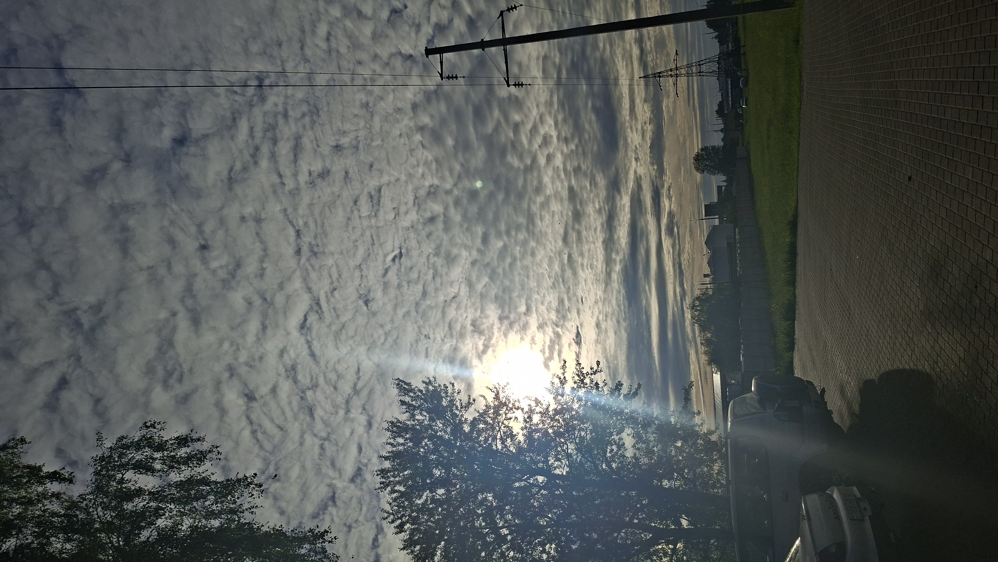 The image depicts an outdoor scene in Smaliavichy, Belarus. A paved area in the foreground is occupied by two parked vehicles: a silver car on the left and a darker SUV on the right, partially obscured by a tree. A large tree with dense foliage stands on the left side of the frame, partially silhouetted against a bright sun. The sun is low in the sky, casting a lens flare and illuminating a cloud-filled sky. A tall power line pole with insulators is visible on the right side of the image, along with distant power lines and other industrial structures. A strip of green grass borders the paved area, behind which a fence runs. The overall impression is a slightly overcast day with the sun breaking through.