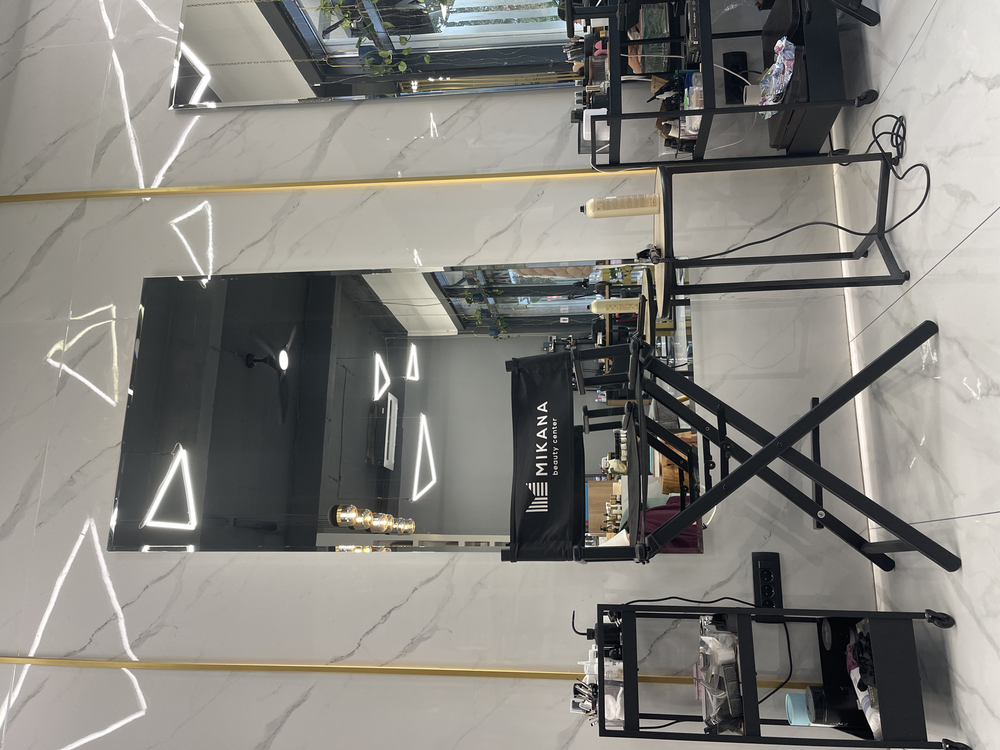 A photograph taken in Budva, Montenegro, showcases a beauty center's interior. The walls feature a marble pattern, and a horizontal gold accent stripe is present. Two rectangular mirrors are mounted on the wall, reflecting lights shaped like triangles and a hanging light fixture with spherical bulbs. A makeup chair is positioned in front of the lower mirror, and a black rolling cart is placed nearby. The mirror also reflects other interior elements, including plants and beauty products.