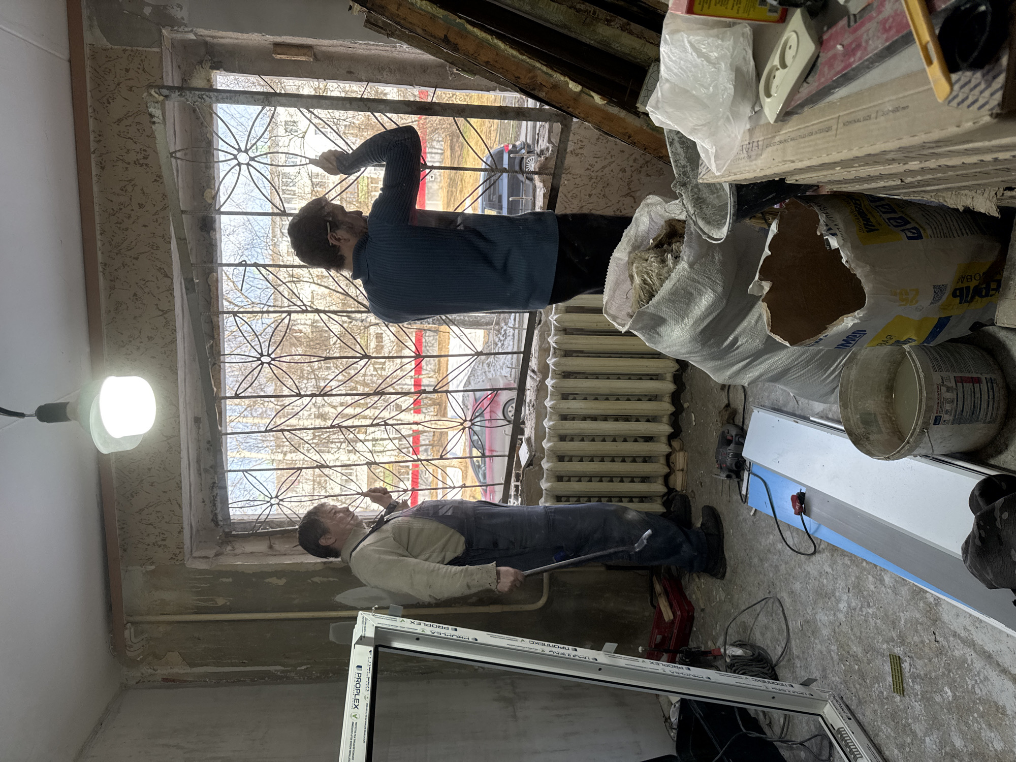Here is a detailed description of the image:
**Scene:** An interior view of a room undergoing renovation.
**Location:** City of Chaikovsky, Russia.
**People:** Two male individuals are present.
* One man is standing, leaning toward the window, seemingly working on the window frame. He is wearing a blue sweater and dark pants.
* The other man is lying on the floor. He is wearing a light-colored shirt and blue overalls, holding a metal bar or tool.
**Subjects and Objects:**
* A large window with a decorative metal grille is present. Through the window, elements of an urban landscape with buildings and vehicles are visible.
* A radiator is located below the window.
* A new white window frame leans against the wall near the man on the floor. The words “ПРОПЛЕКС” and "PROPLEX" are printed on the frame.
* Construction materials, including bags of material, are stacked in the room.
* A light bulb is hanging from the ceiling.
* A bucket, various tiles, and tools are among the construction items in the room.
**Actions and Interactions:**
* The two men are engaged in what appears to be a window replacement or renovation project. The standing man is focused on the upper part of the window, while the man on the floor possibly is installing the new frame.
**Environment:**
* The room appears to be in a state of disrepair, with unfinished walls and debris scattered on the floor, indicating ongoing construction work.