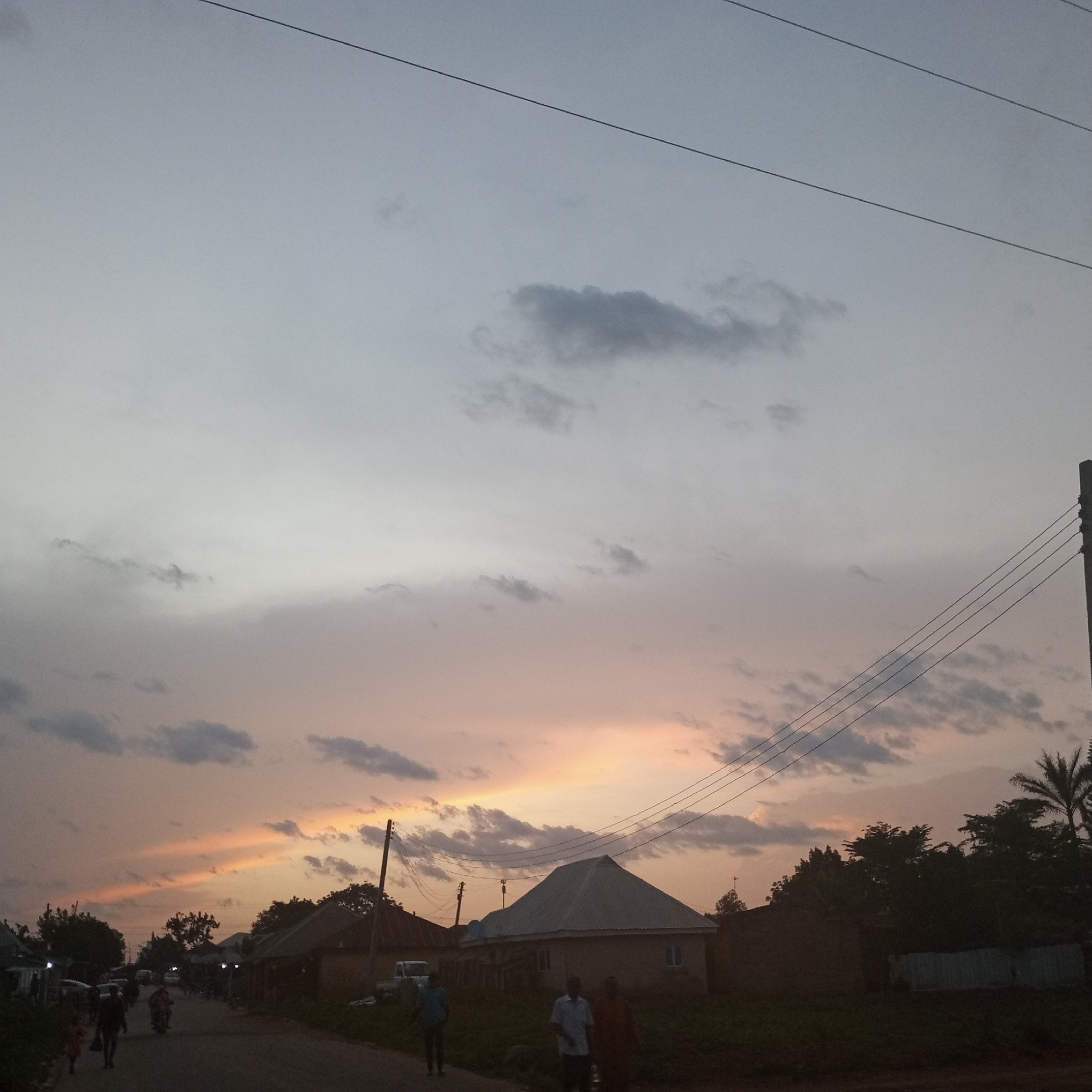 The image depicts a street scene at dusk in Kuru Dungus, Nigeria. The sky is a mix of grey and orange hues from the setting sun, with scattered clouds and visible power lines. A row of low-rise buildings lines the street, with varying roof types. People are walking along the road. A few vehicles are parked or moving in the background. Vegetation is present along the edge of the road and behind the buildings. The overall atmosphere is that of a quiet, rural community at twilight.