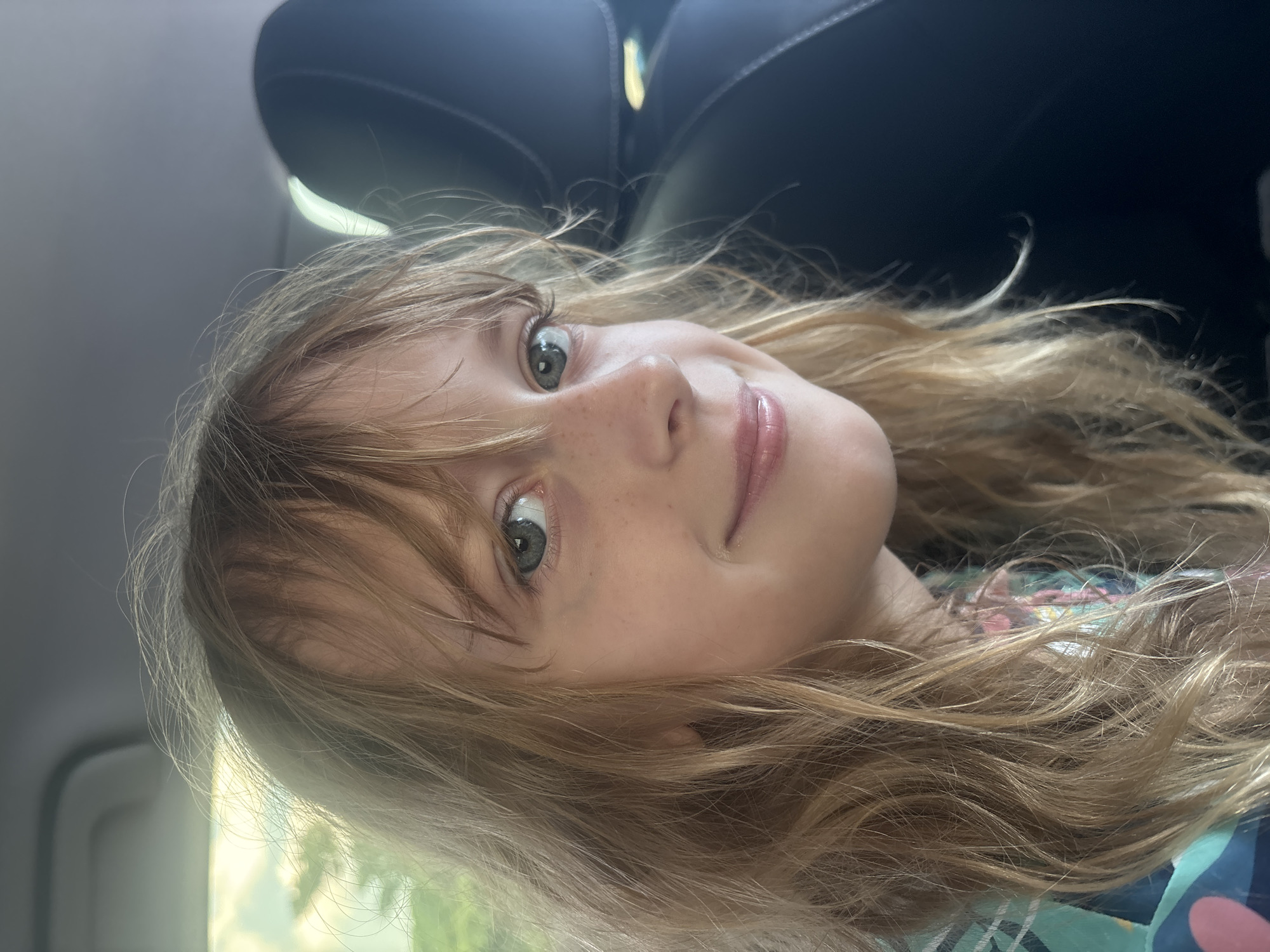 The image shows a close-up, head-and-shoulders view of a young girl with blonde, wavy hair. She has fair skin with freckles, blue eyes, and is smiling gently. She is wearing a colorful, patterned shirt. The background is blurred, suggesting she is in a vehicle. To her right, a dark headrest of a car seat is visible. The lighting suggests it is daytime, with some ambient light coming from the left, possibly through a car window. The image does not contain enough information to determine its location in Liwan District, China.
