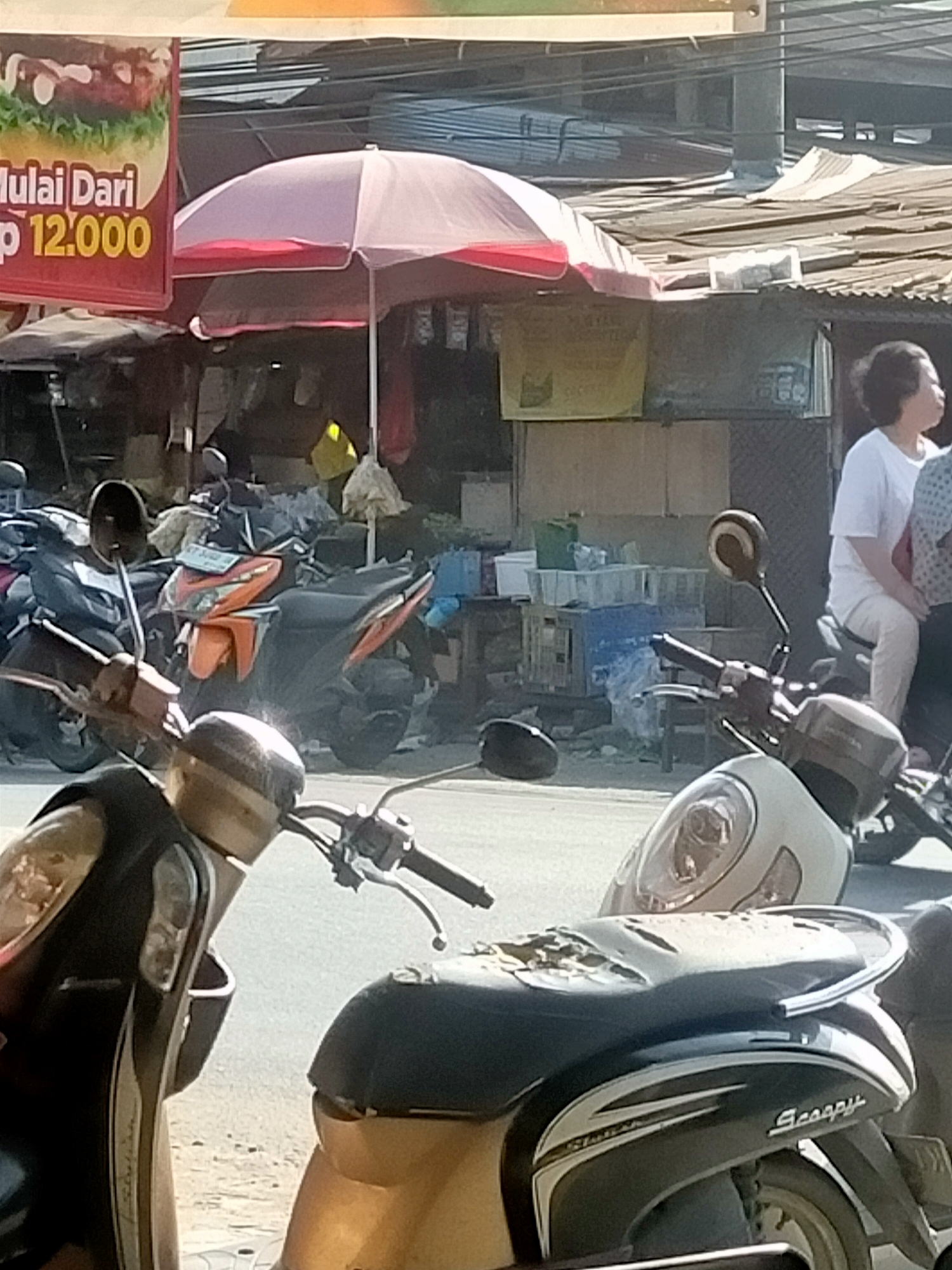 This is a street-level, medium-distance, eye-level shot of a market scene with motorcycles in the foreground. A red sign with white text that reads "Mulai Dari" and yellow text that reads "Rp 12.000" is visible on the left. Behind the sign, a pink and red umbrella shades a market stall. A person in a white shirt and pants is seated on a motorcycle to the right of the frame, looking away. Several other motorcycles are parked in the background, along with market stalls constructed from corrugated metal and wood. The ground is paved, and power lines are visible overhead. The lighting suggests it is daytime.