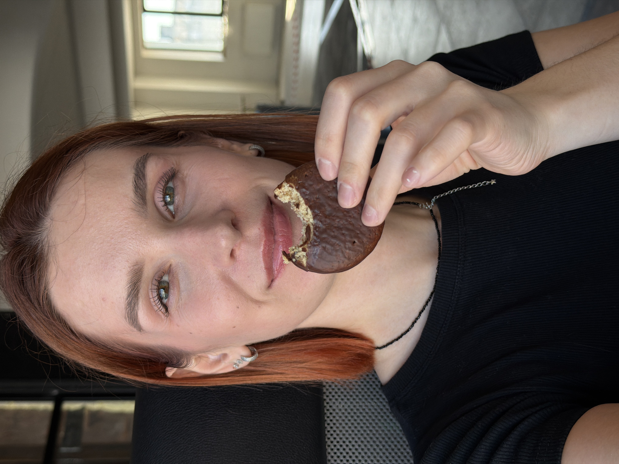 A light-skinned woman with auburn hair is angled to the left, holding a partially eaten chocolate-covered cookie to her mouth. She wears a black ribbed shirt, and a black beaded necklace is visible. A silver chain necklace with a star charm is also visible. Her ear has multiple silver hoop earrings. The background shows a window with natural light, suggesting an indoor setting. The photo appears to have been taken indoors in a city like Chaikovsky, Russia.