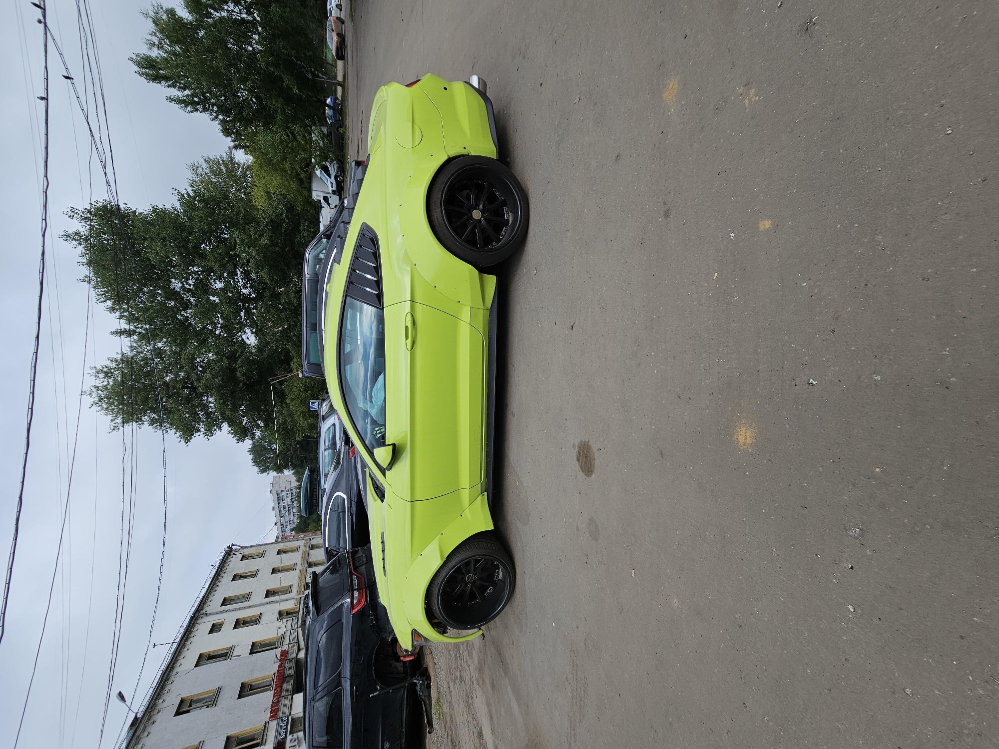 A bright lime green Ford Mustang is parked on an asphalt lot in an urban environment. The car has custom widebody fenders and black aftermarket wheels. In the background, a multi-story building with numerous windows and a row of trees are visible. Several other vehicles are parked in the vicinity. The sky is overcast. The scene is likely located in Moscow, Russia, given the context.