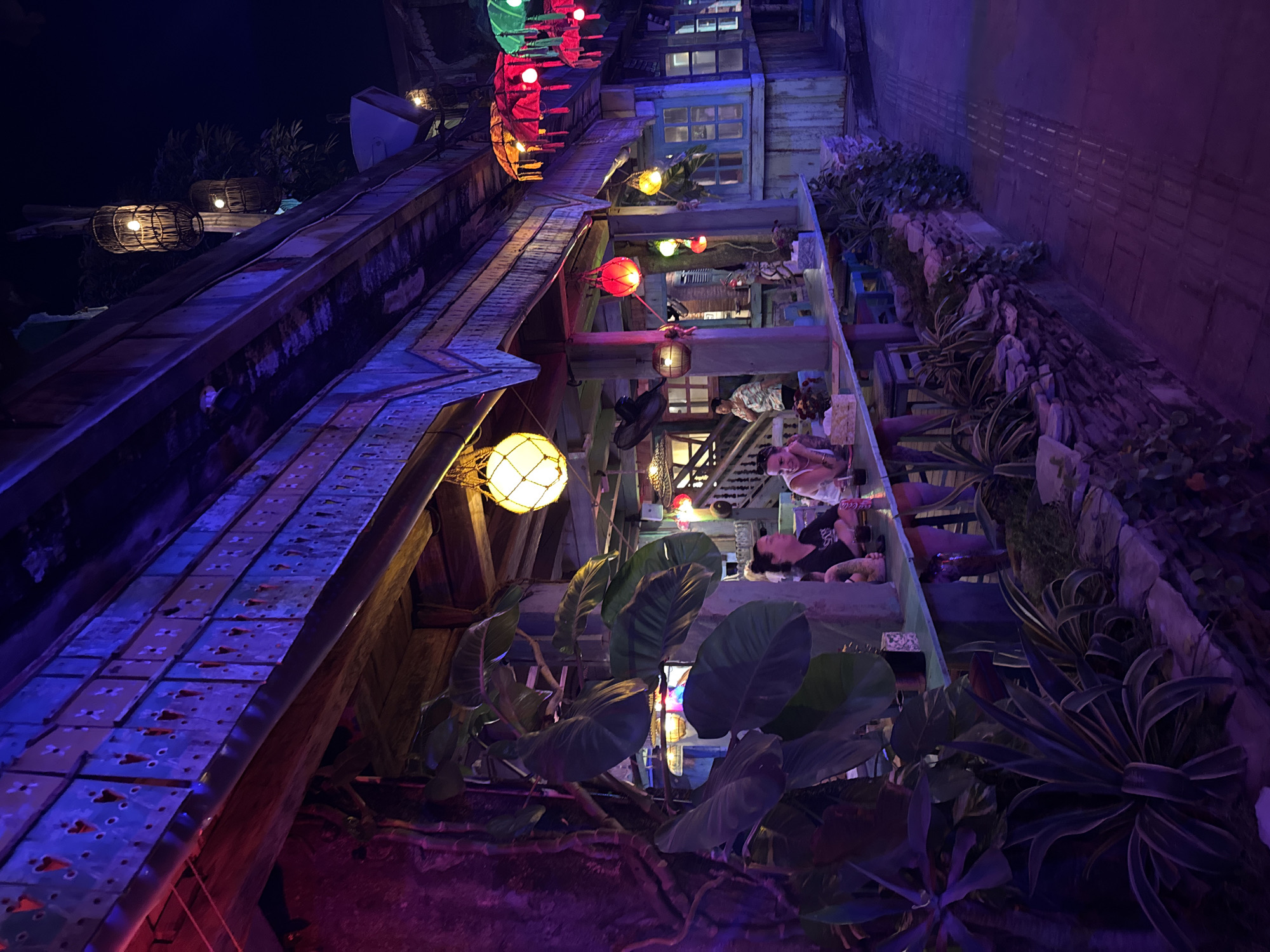 This image depicts a vibrant outdoor dining area at night, likely a restaurant or bar in Kuta, Indonesia. The scene is illuminated by an array of colorful lights, including hanging lanterns and decorative umbrellas in red, green, and yellow. The architecture features a rustic wooden structure with a decorative, patterned fascia board that is lit with purple and blue lights. Several large, leafy plants are prominent in the foreground, adding a tropical feel. Two women are seated at a counter, appearing to be engaged in conversation. Other patrons and staff are visible further inside the establishment, where more windows and structures are apparent. The overall atmosphere is lively and inviting, characteristic of a popular nightlife spot in a tourist destination.