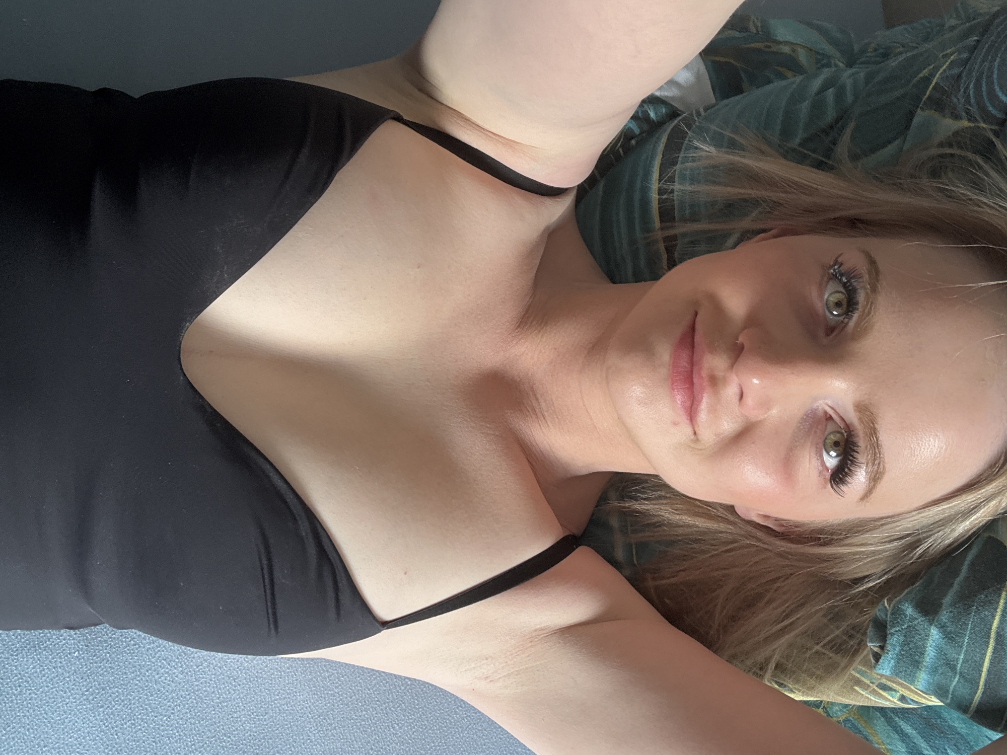 This media file features a fair-skinned individual, likely a woman with blonde hair, lying down and looking directly at the camera. She is wearing a simple black tank top or camisole with thin straps. Her arms are casually raised above her head, creating a relaxed pose.
The subject's head rests on a pillow with a distinctive green and teal striped or swirling pattern. Below her body, a plain light blue or grey surface is visible, suggesting a bed or a soft floor covering. Her eyes are a striking green, accentuated by prominent eyelashes, and her lips are a soft pink. She has a calm expression with a subtle smile, engaging the viewer directly. Her blonde hair is loosely styled, framing her face.
The scene appears to be set indoors, likely a bedroom or living area, with soft, even lighting that does not strongly indicate a specific time of day. There are no visible texts or distinct cultural cues within the image itself to confirm the provided location context of Saint Petersburg, Russia. The overall mood is relaxed and personal.