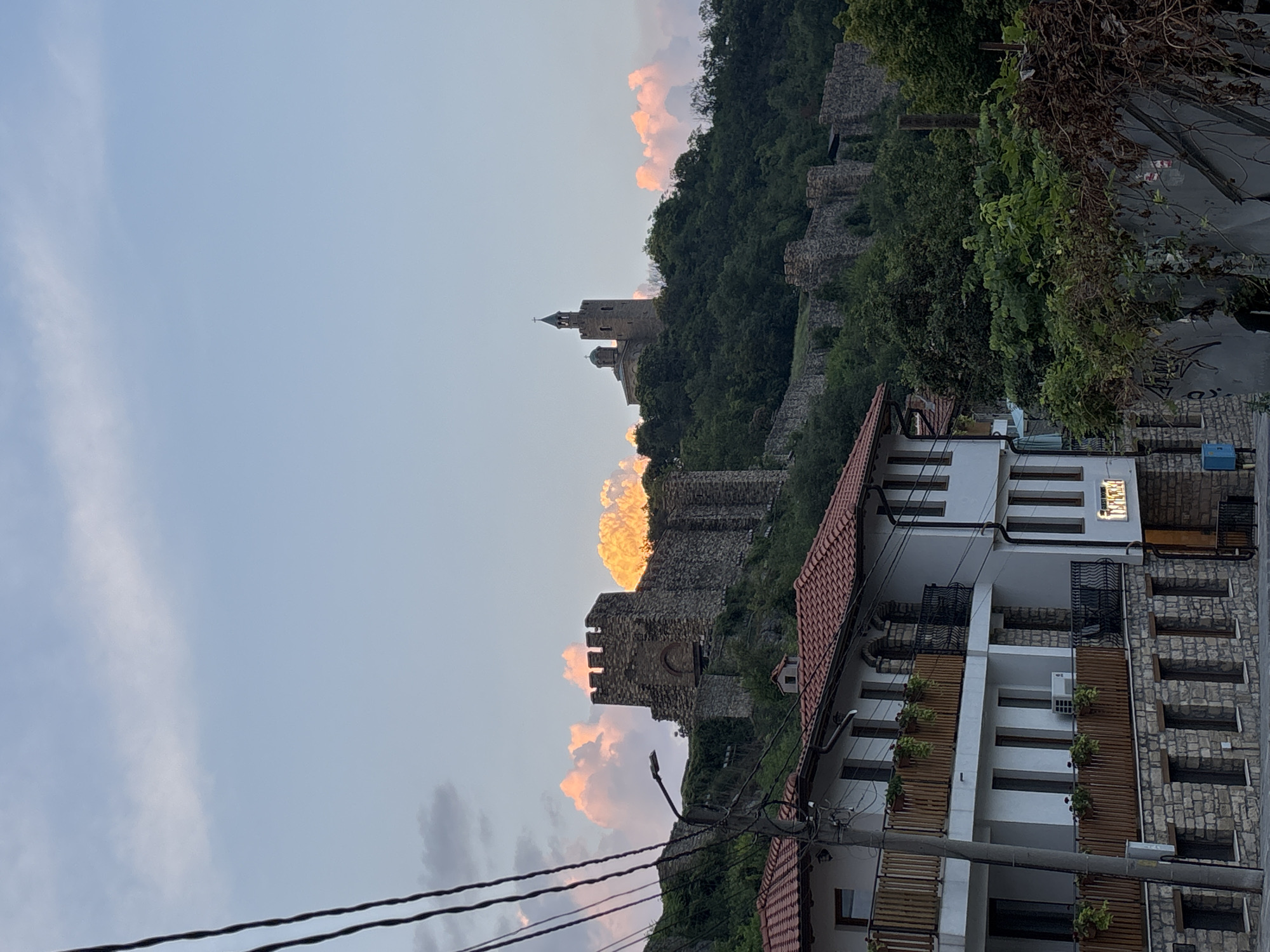 The image shows a view of Tsarevets Fortress in Veliko Tarnovo, Bulgaria, at sunset. The fortress, built of stone, sits atop a hill covered in green trees. The sky is a pale blue with wispy clouds illuminated by the setting sun, casting a warm glow on the fortress and the clouds behind it. In the foreground, there are several multi-story buildings with red tile roofs and balconies. The buildings appear to be a mix of residential and commercial properties. Power lines are visible in the upper left corner of the frame, crossing the sky. The overall scene depicts a historic European town bathed in the soft light of dusk.