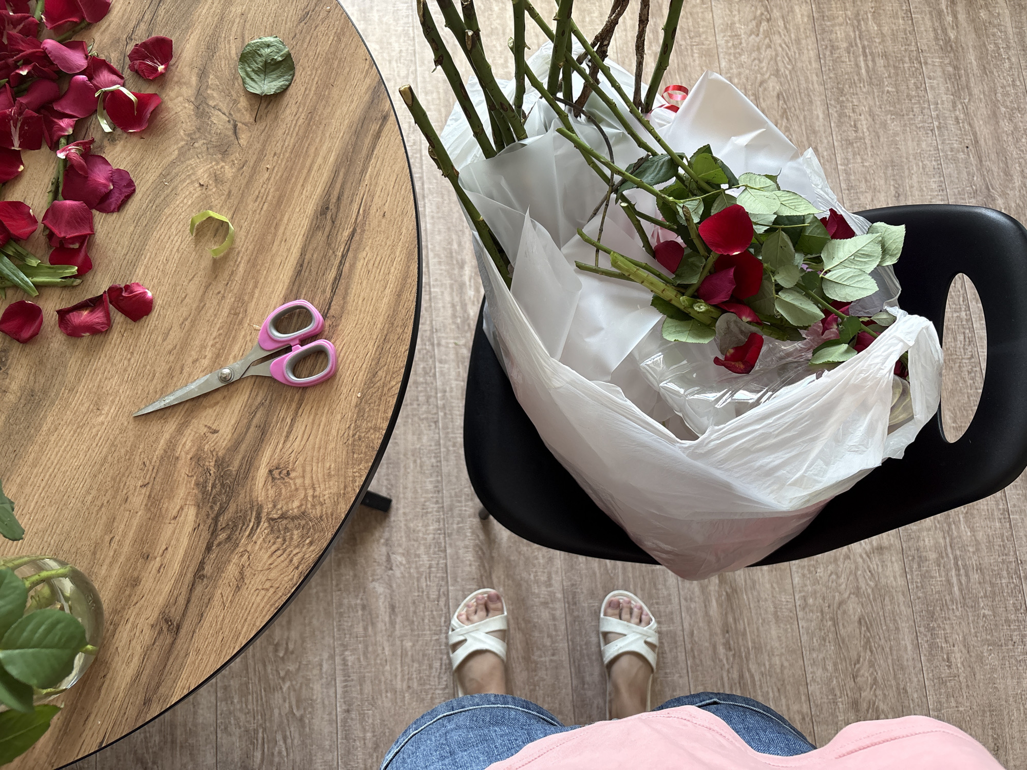 An overhead shot captures a natural wood coffee table on which are scattered red rose petals, a pair of pink-handled scissors, and a single dried green leaf. A glass vase holding a few green rose leaves and stems is visible in the upper left quadrant. In the lower half of the frame, a person's legs are visible from the knees down, wearing blue denim jeans and white sandals, with their feet resting on a light wood floor. Beside the person's feet is a black chair, partially covered by a white plastic bag filled with trimmed rose stems, more red petals, and green leaves. The scene appears to be an indoor domestic setting, likely a living room or dining area. The flooring is consistent with wooden laminate. The image is taken from a high angle, looking directly down. No specific landmarks identify the location as Rostov-on-Don, Russia.