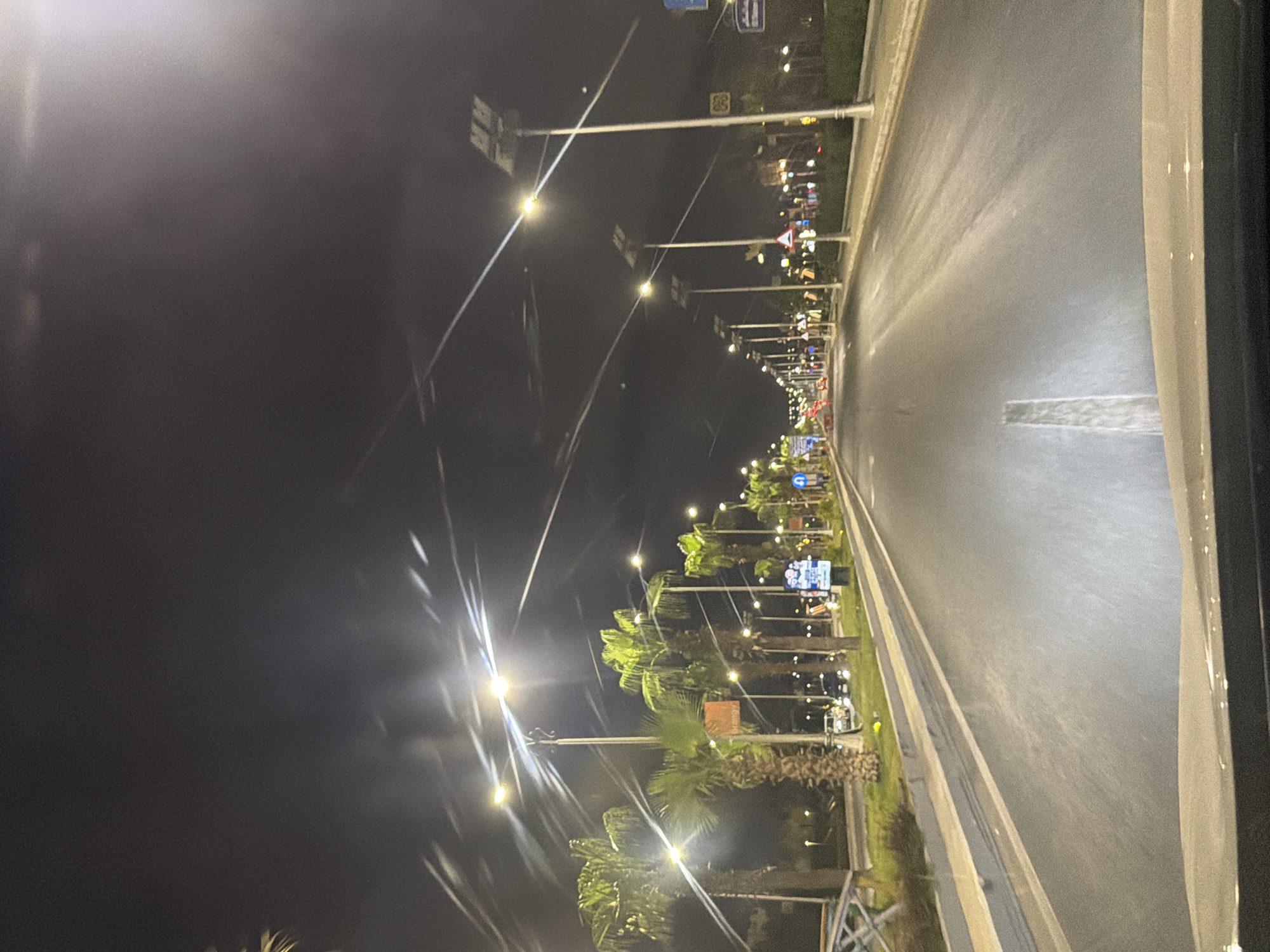 The image depicts a nighttime scene on a wide, paved road in Sharm El Sheikh, Egypt, likely a main boulevard. The road is lined with palm trees and modern streetlights, some equipped with solar panels. On the right side of the road, there are several illuminated signs and a traffic sign indicating a U-turn is permitted. On the left, there is a barrier and some greenery. The road surface appears wet, reflecting the streetlights. Several cars are visible on the road, some in motion. The sky is dark and clear, with the bright lights of the streetlights creating starburst effects in the photograph. The perspective suggests the photo was taken from a moving vehicle.