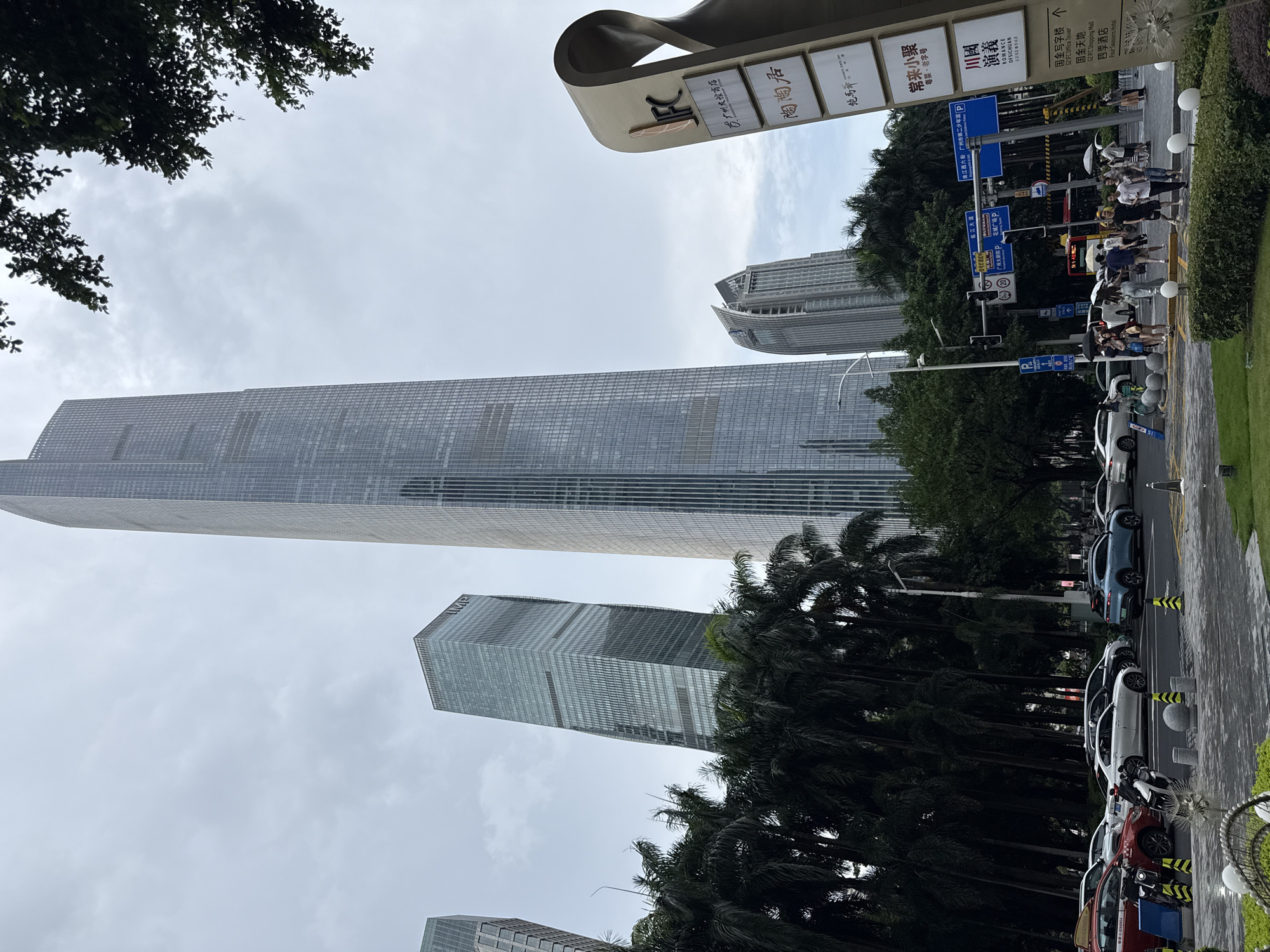 The image shows a street scene in Tianhe District, China, with several tall skyscrapers dominating the background. The IFC (Guangzhou International Finance Center) tower is the most prominent, a slender, glass-facade skyscraper. To its left is another skyscraper, and further to the left, a shorter, block-like building with glass panels. To the right of the IFC, a curved building with a unique architectural design rises.
In the foreground, palm trees line the street, obscuring some of the lower portions of the buildings. A signpost with multiple panels is visible on the right, displaying Chinese characters and the IFC logo, suggesting a commercial area. The street is busy with traffic, including cars, a red taxi, a bus, and motorcycles. Pedestrians with umbrellas are present, indicating overcast or potentially rainy weather. Traffic lights and street signs are visible, directing the flow of vehicles. The ground is paved with asphalt and patterned tiles, with a manicured green lawn and ornamental spheres in the lower right corner. The sky is heavily clouded, creating a diffused light.