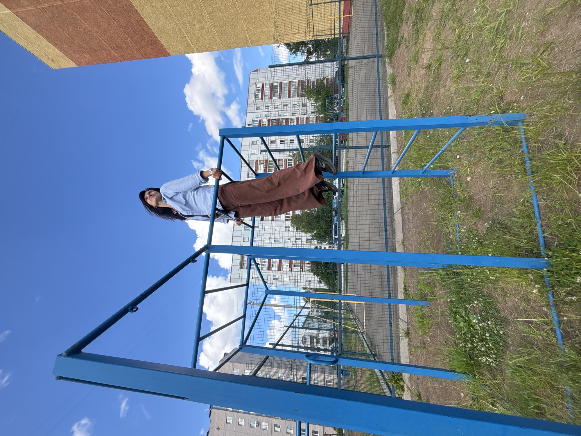 A woman is performing exercises on a blue metal monkey bar frame in a residential area of Chaikovsky, Russia. She is wearing a light blue collared shirt and brown pants, with her hands gripping the bar above her head. Her body is suspended mid-air, and she is looking upwards. The monkey bar structure is set in an outdoor urban environment, with a chain-link fence visible behind it, enclosing a paved area with markings. Beyond the fence, there are several multi-story apartment buildings, typical of Russian residential architecture, under a bright blue sky with scattered white clouds. Green grass and some small white flowers grow around the base of the monkey bars. The overall scene suggests a sunny day in a public outdoor recreational space within a city.