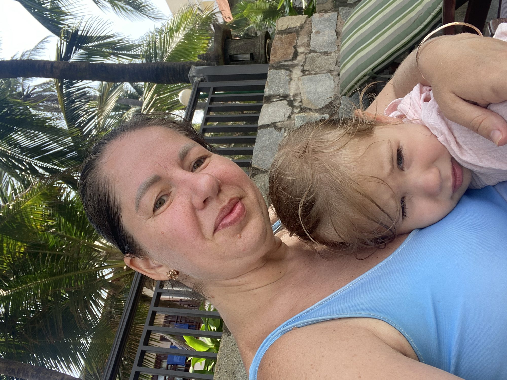 This is a selfie of two people, a woman and a baby, likely taken outdoors in Phu Thuy Ward, Vietnam. The woman is in the foreground, looking towards the camera with a slight smile. She has dark hair and is wearing a light blue top. The baby is nestled against her, with their face turned away from the camera and eyes closed, appearing to be asleep or resting. The baby is wrapped in a light pink fabric.
In the background, there are palm trees and a stone wall, suggesting a tropical or sub-tropical environment. A striped cushion is visible on what appears to be outdoor furniture. The overall lighting suggests it is daytime, possibly late morning or early afternoon, with bright, diffused sunlight filtering through the palm fronds. The scene evokes a relaxed and intimate atmosphere, possibly during a vacation or a moment of quiet time together. No text is visible in the image.