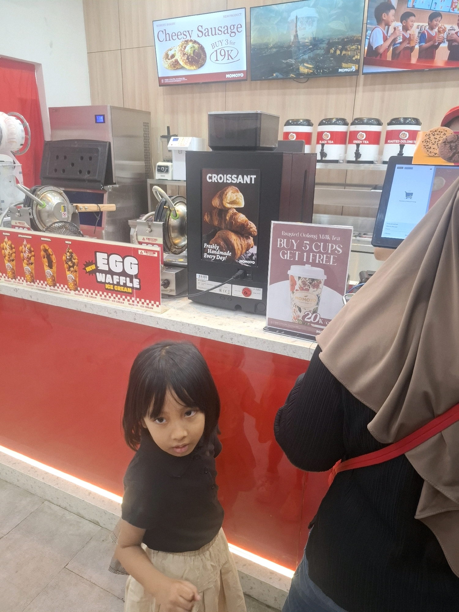 In Cianjur, Indonesia, a young girl with dark hair and a black shirt stands on a tiled floor in front of a vibrant red counter. Her skirt is a light tan color. Behind her, a modern food service counter is visible, with various promotional displays and equipment. One screen advertises "Cheesy Sausage," while another features "Croissant" with an image of the pastry. A third display promotes "Roasted Oolong Milk Tea" with a "Buy 5 Cups, Get 1 Free" offer. Several dispensers for drinks, labeled "Black Tea," "Green Tea," and "Roasted Oolong Tea," are lined up on a shelf. To the left, a sign for "Egg Waffle Ice Cream" shows different flavors. The overall environment suggests a fast-food or dessert shop.