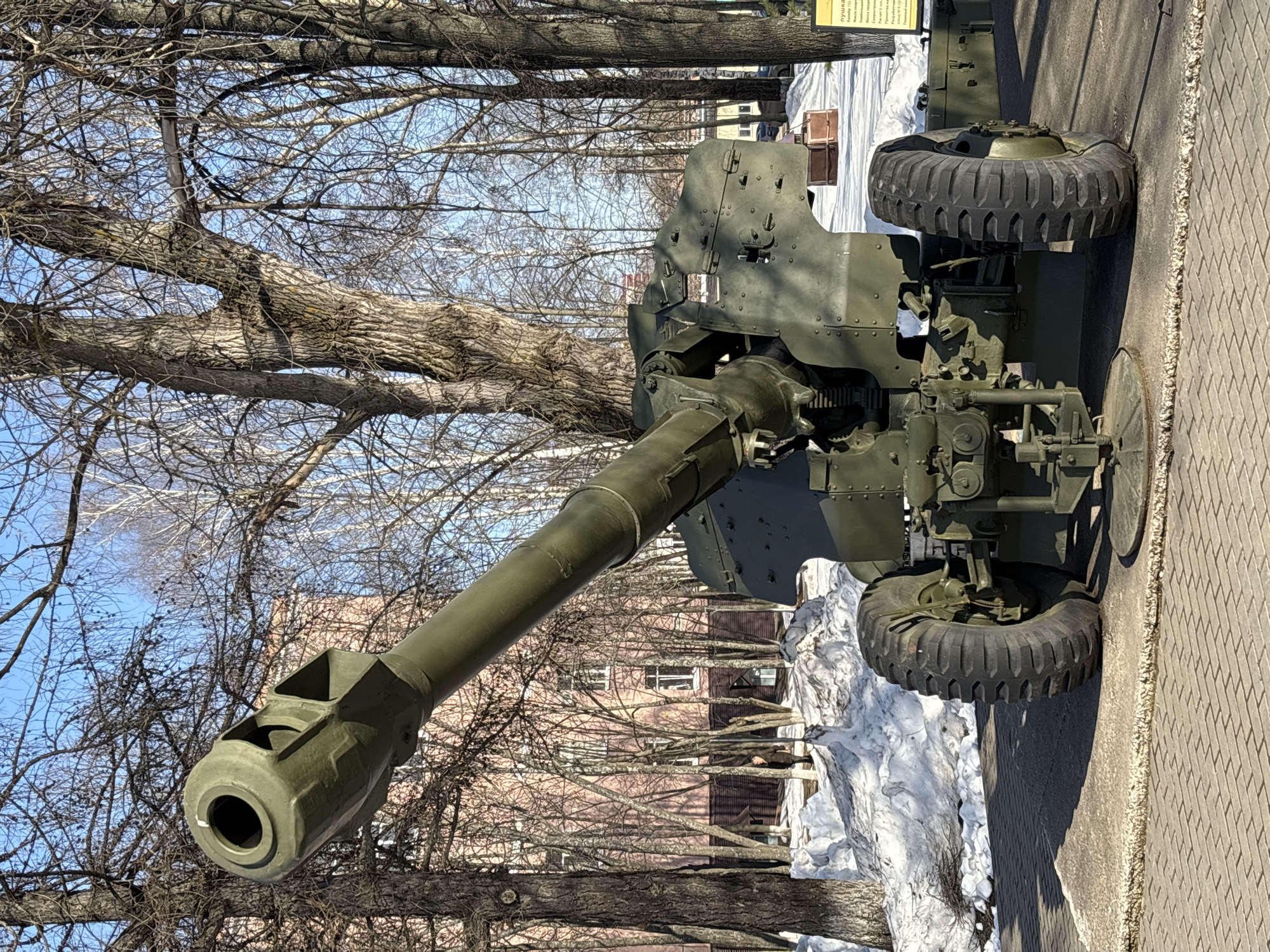 A Soviet 122 mm howitzer M-30 of WWII vintage stands in an outdoor environment on a paved surface in Izhevsk, Russia. Trees are visible in the background, as is a building, with snow present on the ground. The howitzer is green and has two tires.