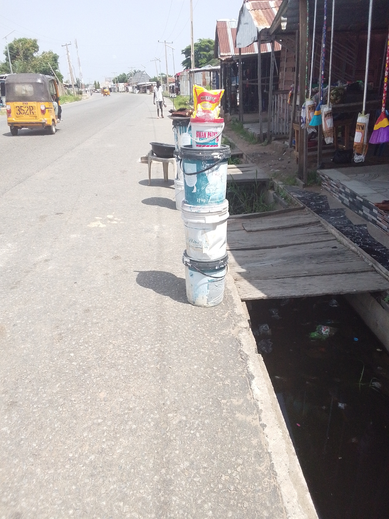 This image depicts a street scene in Jalingo, Nigeria, likely during the daytime under clear, sunny weather. The foreground is dominated by a paved road and a concrete drainage channel running alongside it. Several plastic buckets are stacked precariously on the edge of the drainage channel. The buckets are of various sizes and colors, with some appearing to contain liquids or other substances. One bucket has "SWAN PAINT" printed on it, and another has "27 kg".
A yellow motorized tricycle, commonly known as a "keke napep," with the license plate "352R" is visible in the middle ground, driving down the road. A person is seated behind the driver. Further down the road, a man in a white shirt and dark trousers walks away from the camera.
On the right side of the image, there are several makeshift stalls with corrugated metal roofs. Colorful brooms hang from the front of one of the stalls. A yellow bag, possibly containing food items, is placed on top of a bucket in front of the stalls. The overall setting suggests a market or commercial area along a busy street. The drainage channel appears to contain murky water with visible litter, indicating potential environmental concerns. The scene evokes a sense of everyday life and commerce in a Nigerian town.