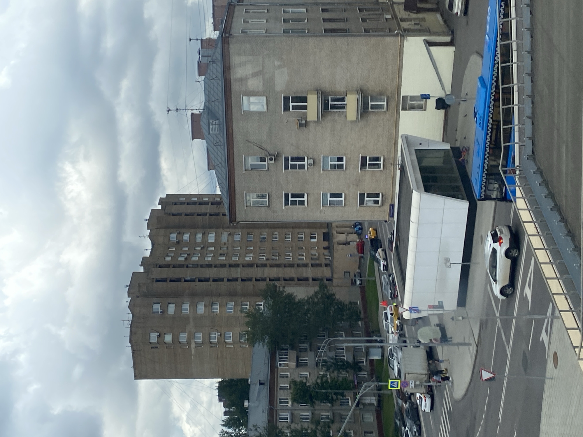 The image depicts a cloudy day in Moscow, Russia, focusing on a street intersection and surrounding buildings. A large, tan-colored, multi-story apartment building with many windows is prominent in the background, to the left. To its right, a grey, brick apartment building with a flat, dark roof and visible air conditioning units on its facade is in the mid-ground. A modern, white, angular structure with large glass windows, possibly a kiosk or small building, is situated at the intersection. Several vehicles are present, including a white SUV, a police car, a yellow taxi, a truck, and a blue bus with white decorative trim along its roof. The street is paved, with crosswalks and traffic signs visible. A few people are standing near the intersection. The sky is overcast with heavy clouds.