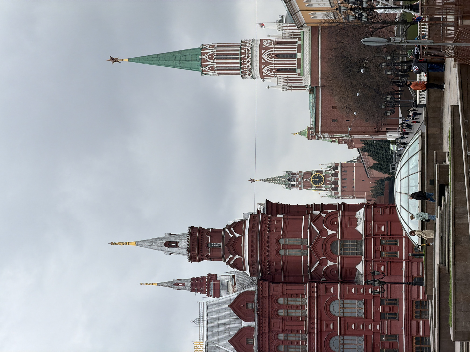 This image captures a vibrant urban scene in Moscow, Russia, showcasing iconic historical architecture under an overcast sky.
In the left and central foreground, the State Historical Museum dominates, characterized by its deep red brick facade, intricate white decorative elements, and multiple spires, some of which are topped with golden finials. Behind and to the right of the museum, the distinguished walls and towers of the Moscow Kremlin are visible. A prominent Kremlin tower features a tall, pointed green spire crowned with a bright red star. Further in the background, towards the right, the Spasskaya Tower is identifiable by its large clock face displaying Roman numerals, also topped with a star.
The right foreground depicts a lively pedestrian area or plaza where several people are engaged in casual activity. Individuals are seen walking and standing on the paved ground, with one person in a distinctive orange jacket particularly noticeable. Modern urban features such as street lamps, railings, and a large, curved glass structure—possibly a skylight or part of a building entrance—are present.
The sky is uniformly light grey and overcast, suggesting a cool, muted daylight. No specific event or particular emotion is discernible; the scene appears to capture typical daily life and tourism in this historic area. Beyond the Roman numerals on the Spasskaya Tower clock, no other clear, readable text is visible.