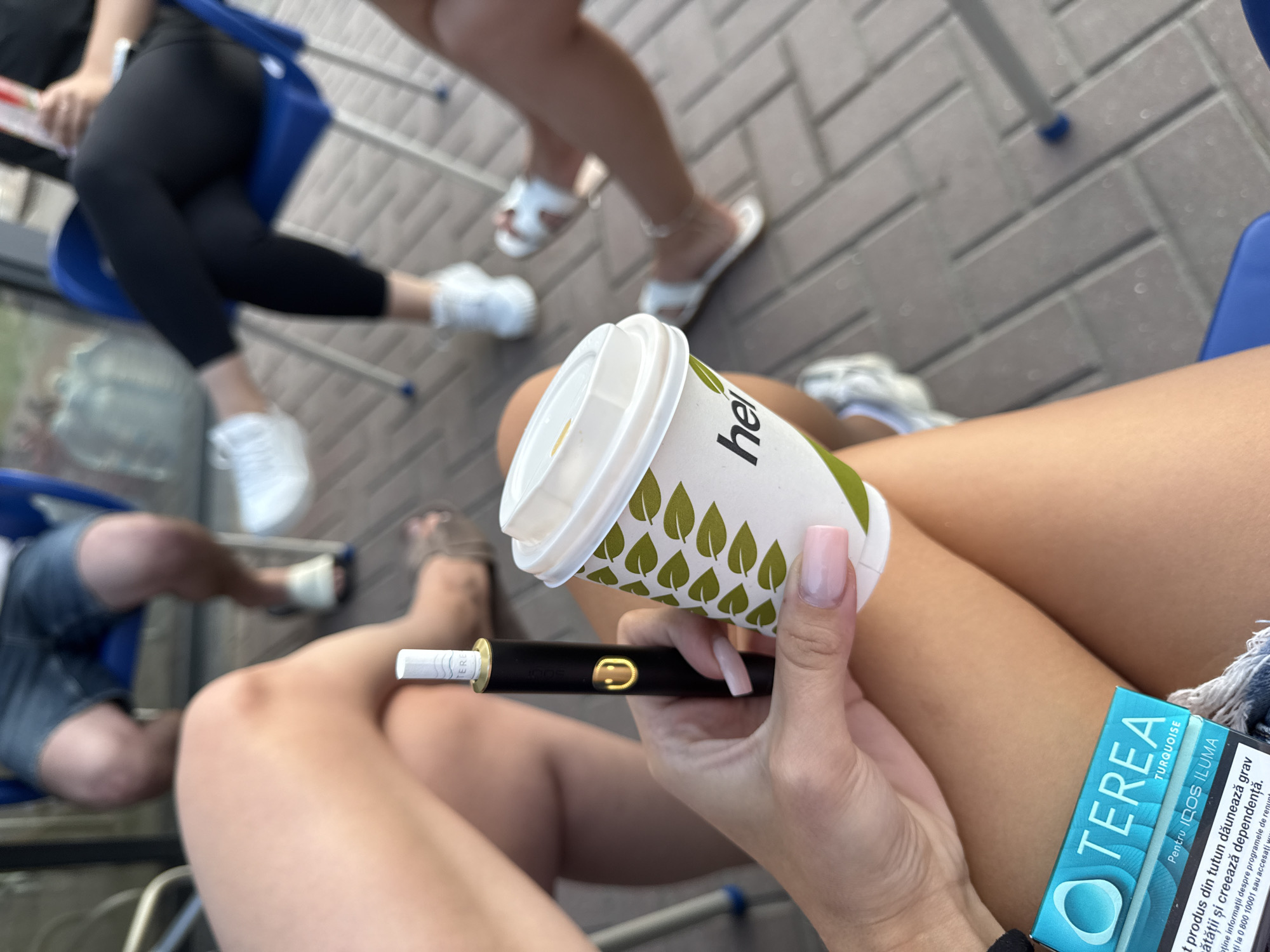 The image shows a close-up of a person's hands holding a black device with a gold smiley face emblem and a white paper cup with green leaf patterns. The device appears to be an electronic cigarette or similar smoking apparatus. The cup has "hel" printed on it. To the left of the cup, a package of "TEREA TURQUOISE" for "IQOS ILUMA" is visible, with Romanian text warning about the harmfulness of smoking. The background is blurred, but shows people sitting in blue chairs, suggesting an outdoor setting like a cafe or public area. The ground appears to be paved with brick-like tiles. The overall scene suggests a casual moment of relaxation or socializing. The location of Brătianovca, Moldova is provided as context for this scene.