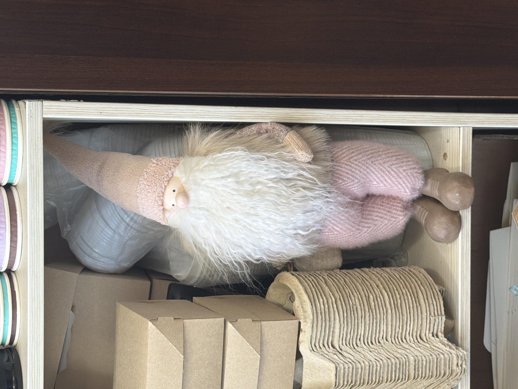 A stuffed gnome doll with a pink knitted hat, white beard, pink patterned pants, and brown shoes rests in a drawer. Other items in the drawer include packing materials, cardboard boxes, and a roll of corrugated cardboard. The drawer appears to be part of a wooden cabinet or shelving unit. The location is not identifiable from the image.