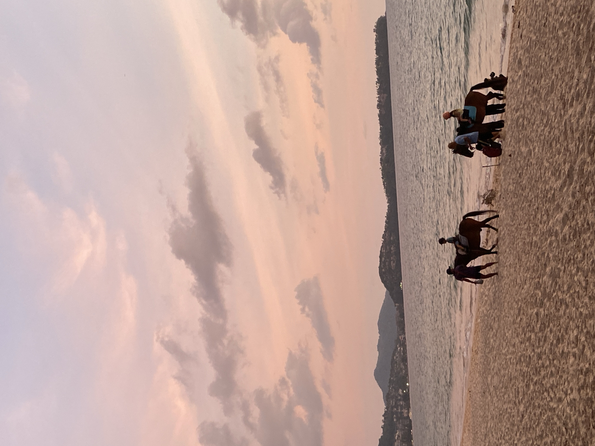 The image captures a serene beach scene at twilight, likely sunset, in Choeng Thale, Thailand. The sky, occupying the upper portion, is painted with soft, diffused clouds displaying hues of light pink, orange, and subtle purple, indicating the transitional lighting of dawn or dusk.
The foreground and mid-ground feature a sandy beach meeting the calm ocean. Gentle waves are visible along the shoreline. Several dark-colored horses are present on the wet sand near the water's edge. In one group, two horses are seen, with at least one person mounted and another person standing beside them, possibly guiding. Further along the beach, another horse is present with a rider and an accompanying person standing nearby, facing the water. The activity appears to be recreational horse riding or a guided beach tour.
In the distance, across the expanse of water, a landmass with rolling hills or mountains is visible, dotted with scattered lights that suggest coastal habitation. The overall ambiance is peaceful and tranquil, showcasing a popular beach activity against a beautiful, colorful sky. No text is visible in the media file.