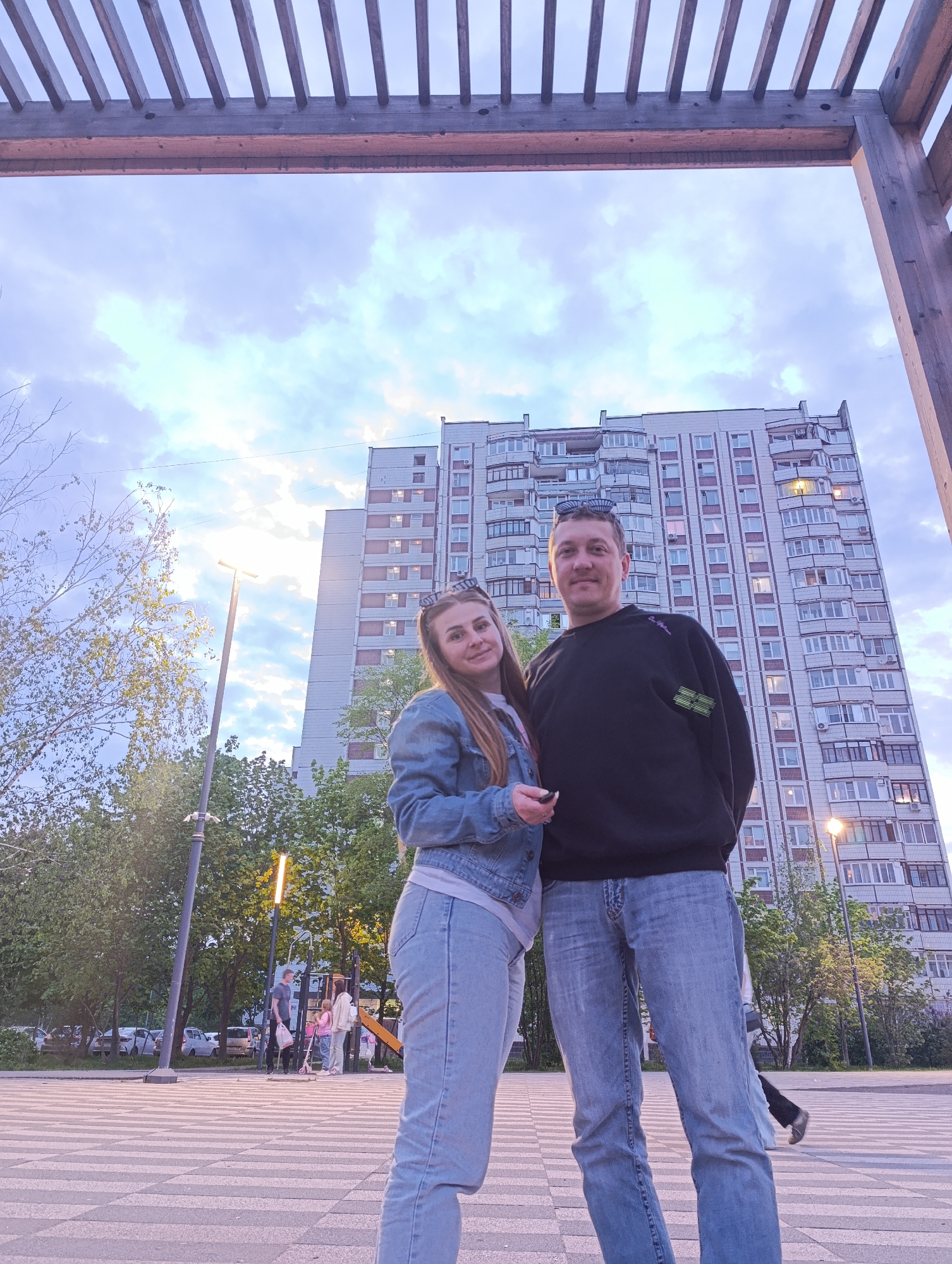 A medium shot depicts a man and a woman standing close together in what appears to be a city park in Unknown, Unknown. The woman, on the left, is wearing a light blue denim jacket and jeans, while the man, on the right, is wearing a black sweatshirt and jeans. Both have sunglasses perched atop their heads. In the background, a tall apartment building is visible, with numerous windows and balconies. The park setting includes trees, street lamps, and a playground area with people in the distance. A wooden pergola frame partially obscures the sky at the top of the image. The ground is paved with a patterned surface. The lighting suggests it is late afternoon or early evening.