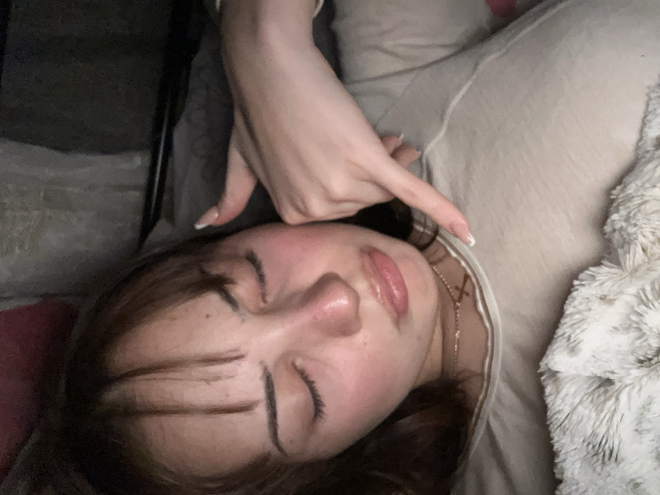 A young woman reclines with eyes closed. She has a fair complexion, dark eyebrows, and brown hair with bangs. Her lips are full and shiny. A hand with manicured nails rests above her head, pointing towards the top right corner. She wears a light-colored top and a delicate gold necklace with a cross pendant. The background includes a soft blanket and part of a dark chair. The overall impression is casual and relaxed.