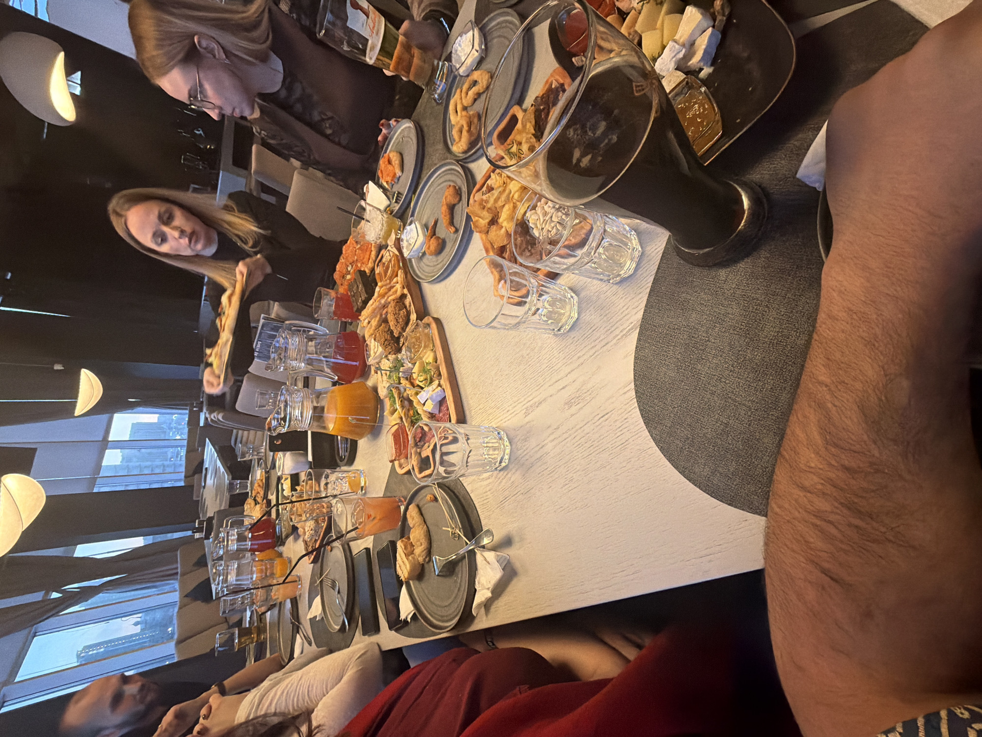 This media file captures a lively social gathering taking place indoors, likely at a modern restaurant or bar, with a potential location in Krasnoyarsk, Russia, given the provided context. The perspective is from someone seated at the table, looking across a spread of food and drinks.
The primary subjects are several people gathered around a large, light-colored table. In the mid-ground, a blonde woman in a dark top is engrossed in her meal, possibly serving food from a platter. Further back, another blonde woman wearing glasses is visible. In the immediate foreground, the hairy arm of a person, likely male, is prominent on the right. Other individuals are partially visible or out of focus around the table, indicating a group setting.
The table is densely covered with a variety of appetizers, snacks, and beverages. Food items include platters with assorted cheeses and cured meats, bread, fried components such as onion rings, chicken pieces, and potato chips, along with various dips. The drinks consist of multiple pitchers of different-colored juices (orange, red, yellow), several cocktails in glasses, and a large glass of dark beer. Plates, empty glasses, and cutlery are also scattered across the table.
The setting features modern decor with dark walls and distinctive, dome-shaped light fixtures. Large windows in the background reveal an urban cityscape with tall buildings, suggesting an evening or night setting due to the reliance on artificial lighting and the appearance of the sky outside. The overall activity is a casual dining or drinking event, implying friends or colleagues enjoying each other's company over food and drinks. The atmosphere appears relaxed and social. No discernible text is clearly visible on any objects within the image.