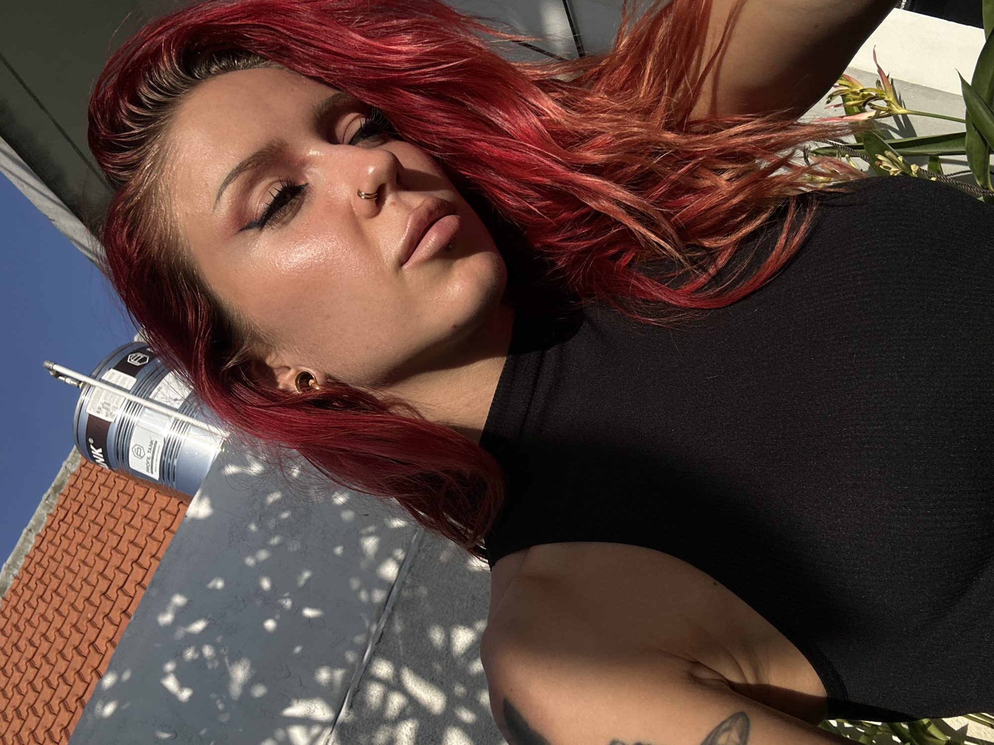 A close-up, eye-level, outdoor medium shot captures a woman with vibrant red hair, her head tilted down, with her eyes closed. The subject is wearing a black tank top and subtle eye makeup with eyeliner. She has a gold hoop earring in her left ear and a double-hooped nose ring. Sunlight illuminates her face, creating a dewy sheen. In the background, a textured, white wall with dappled shadows suggests an outdoor setting. A tiled roof and a large metal water tank are visible in the upper left corner of the image. Green foliage and plants are present in the lower right. The setting appears to be a residential area, likely a rooftop or balcony, in North Kuta, Indonesia, given the architectural style and climate.