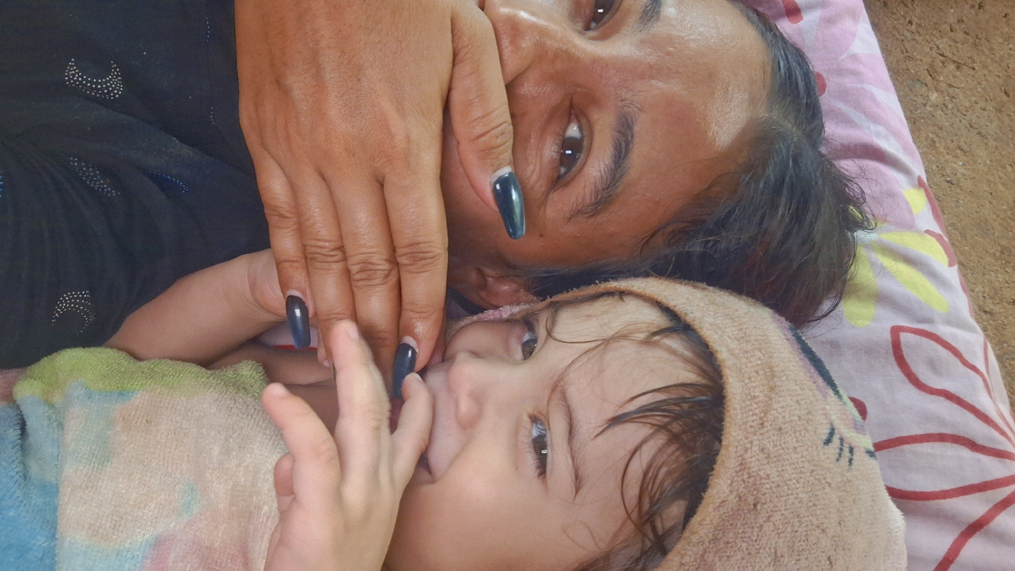A high-angle close-up photograph captures an adult female and a young child in an indoor setting in Vallecito, Venezuela. The adult, positioned on the left side of the frame, has a medium-dark complexion, dark hair, and brown eyes, with visible creasing around the eyes indicative of a smile or laugh. Her right hand, adorned with long, pointed, dark-colored artificial nails, covers her mouth. She is wearing a dark garment with scattered white or silver patterns, including crescent moon shapes and dots. The child, situated to the right and partially obscured by the adult's head and shoulder, has fair skin, brown eyes, and light brown hair. The child wears a light beige or tan textured head covering, possibly a towel or hat, with faint dark markings on it. The child's right hand is positioned near their mouth, mimicking the adult's gesture. A multi-colored soft fabric, exhibiting shades of green, blue, and white, is visible beneath the child. Both individuals are looking towards the camera. The background features a pink fabric with red and yellow floral patterns, likely a bedsheet or pillowcase, above which is a light brown, coarse-textured surface, possibly a wall or floor.