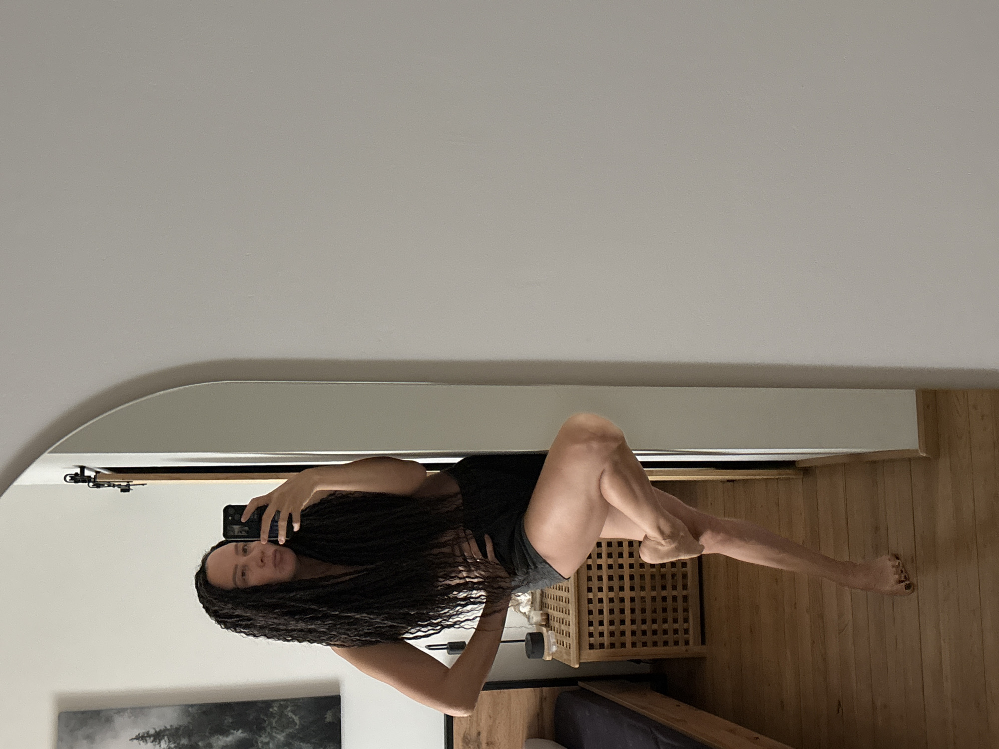 An indoor full-body mirror selfie shows a woman with long, dark, braided hair. She is wearing a dark top and dark shorts, posing with one leg bent at the knee and the other extended. Her reflection captures her holding a smartphone to take the picture, with a neutral to slightly smiling expression. The setting appears to be a bedroom in a residence in Chaikovsky, Russia, characterized by light-colored walls and a wooden floor.
In the background of the reflection, a wooden bed frame is visible, along with a slatted wooden nightstand or chest. A framed picture depicting a landscape with trees hangs on the wall, partially reflected in the mirror. The lighting is bright and even, suggesting it is daytime. No specific weather conditions are discernible, and there is no visible text.