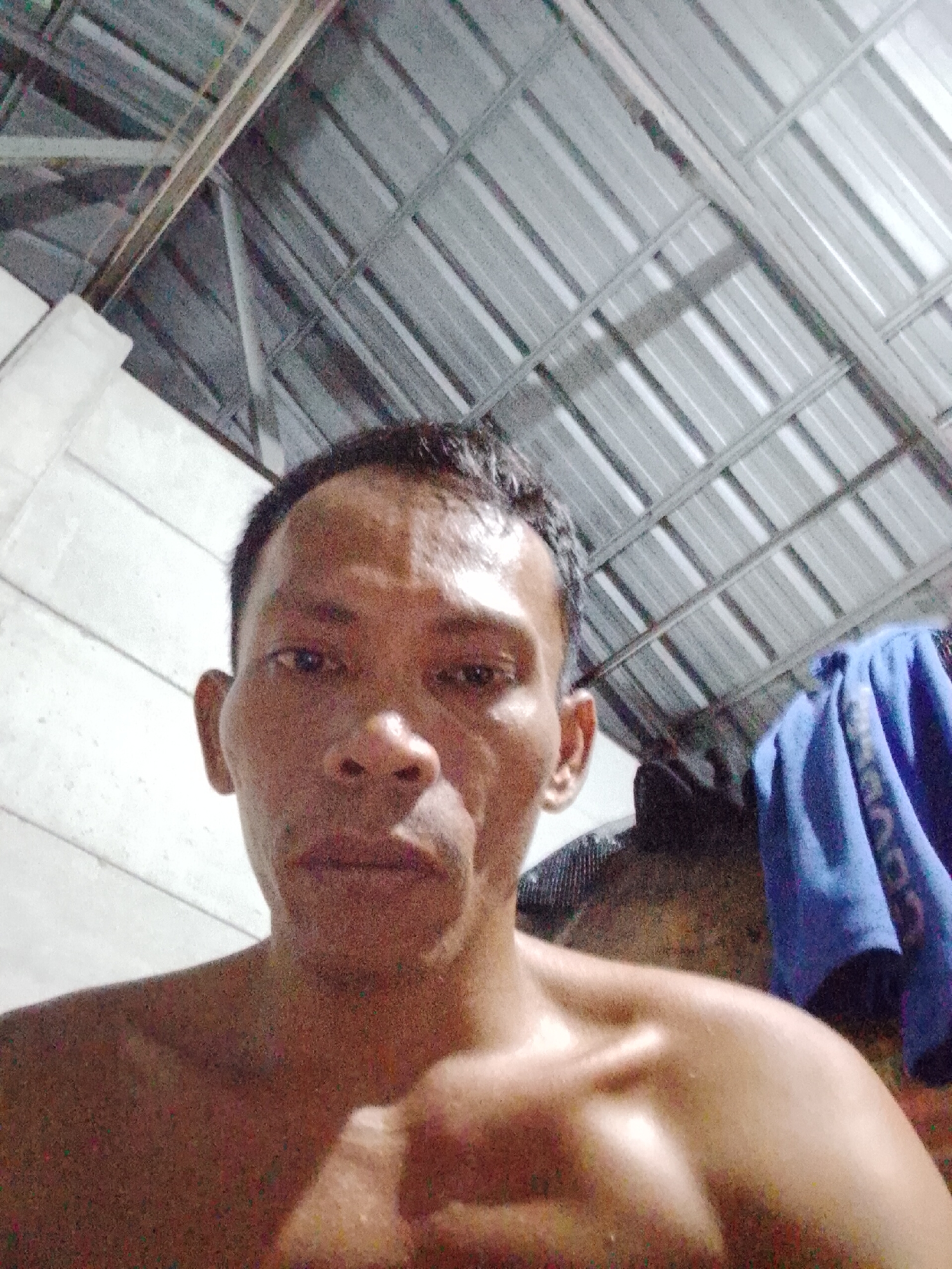 A medium shot shows a man with short black hair, medium brown skin, and a bare chest. He is looking directly at the camera. Behind him is a metal roof and a white wall. A blue cloth is hanging in the background. The image was taken indoors in Bukit Ketok, Indonesia.