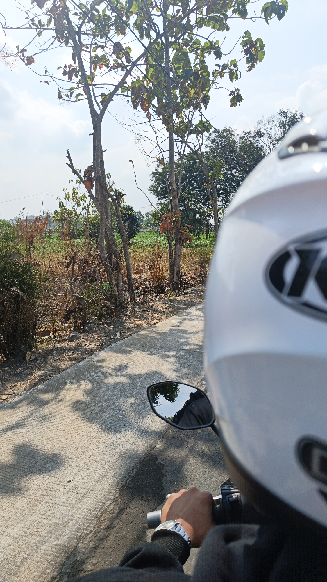 The image is a first-person perspective shot from a motorcycle in Desa Sobokerto, Indonesia. The rider's gloved hand, wearing a watch, is visible gripping the handlebar. A white helmet with a black logo is in the foreground, partially obscuring the view. The motorcycle is on a paved road that curves to the left. To the right of the road, there is dry, scrubby vegetation and several trees, some with brown leaves. Further in the background, there are green fields and more trees. The sky is visible with some clouds, and shadows from trees are cast on the road. The overall scene suggests a rural or semi-rural environment on a sunny day.