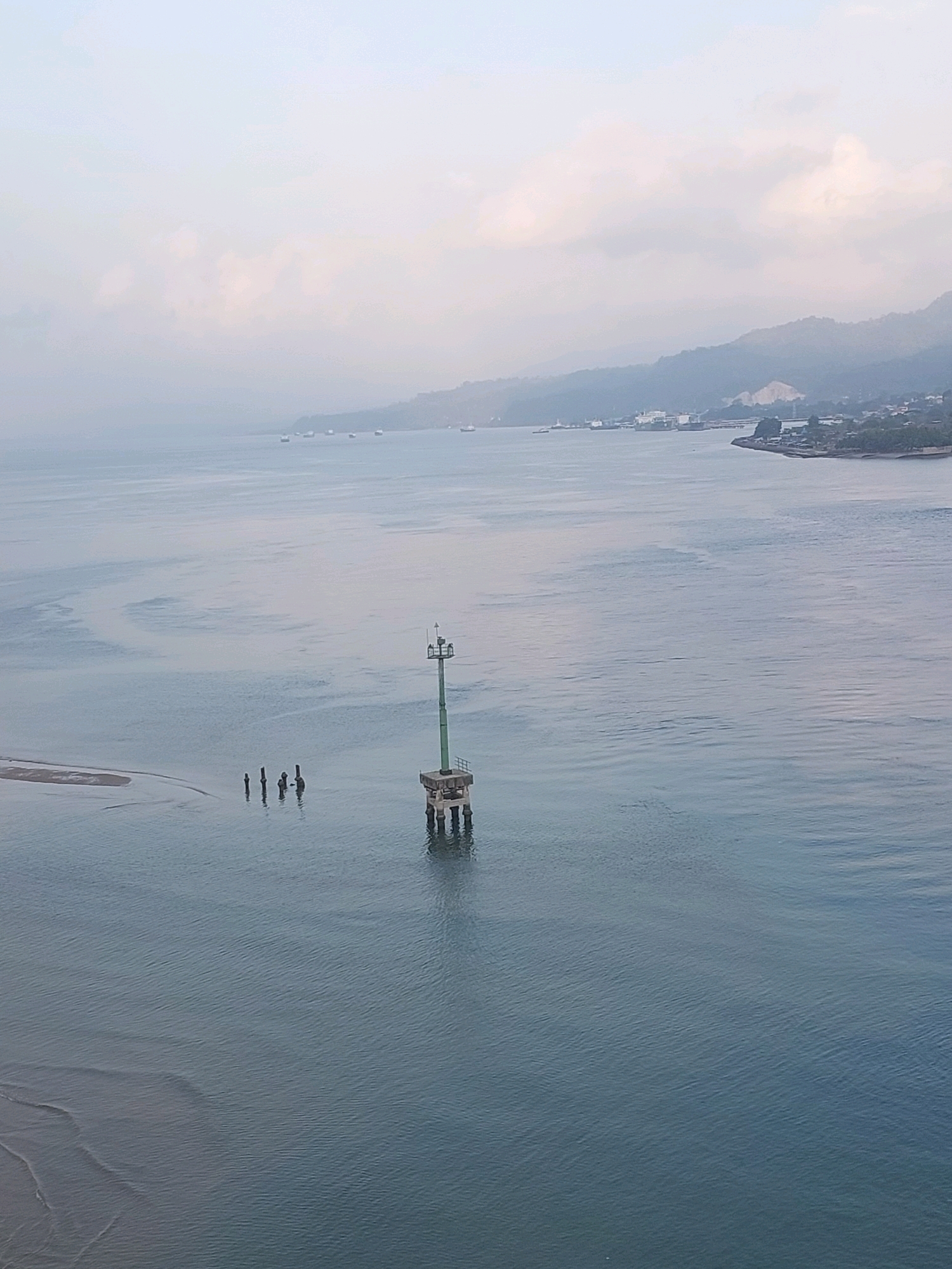 A wide shot captures a body of water with a landmass in the background, likely Ambon, Indonesia. A navigational marker with a green top and concrete base stands in the water to the right of center. To the left of the marker, several dark, slender pilings emerge from the water. The water is calm with gentle ripples, reflecting the pale, overcast sky. Distant boats are visible on the horizon, and the landmass features hilly terrain with scattered buildings and structures, suggesting a coastal town or city. The overall atmosphere is serene and somewhat hazy, typical of a humid tropical climate.