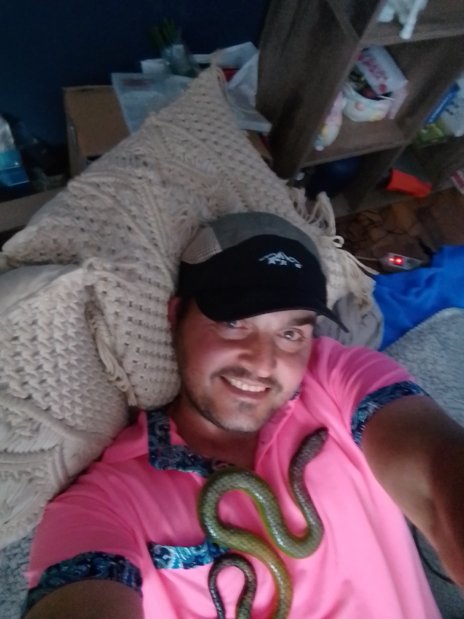 A man wearing a pink polo shirt and a black baseball cap with a white mountain logo is lying down, with a green snake coiled around his neck and upper chest. He is smiling at the camera. The man appears to be indoors, in what looks like a living room. There are shelves in the background filled with various items, and a blue blanket is visible. The lighting suggests it is nighttime or the room is dimly lit.