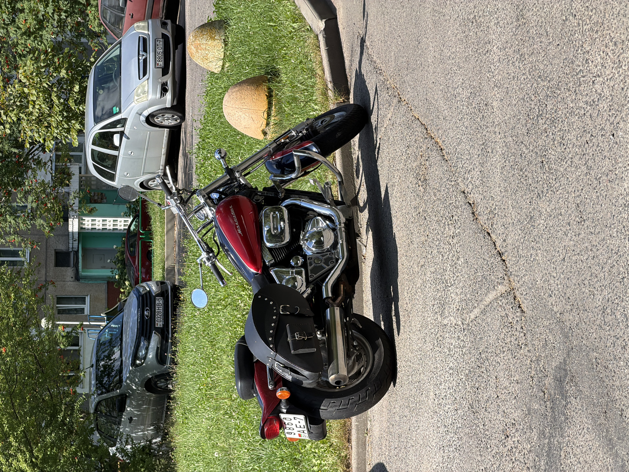 A red and black Suzuki Marauder motorcycle is parked on an asphalt surface next to a curb with a grassy area behind it. The motorcycle has a black leather saddlebag on its right side and a license plate reading "9860 AE-7". In the background, there are two silver cars, one with a license plate reading "8805 OT-7" and another that appears to be a Lada Granta with a license plate reading "0328 HB-5". Behind the cars is a multi-story apartment building with trees and bushes. The scene appears to be a residential area in the city of Minsk, Belarus, judging by the car license plates and architectural style. The lighting suggests it is daytime and sunny. There are no people visible in the image.