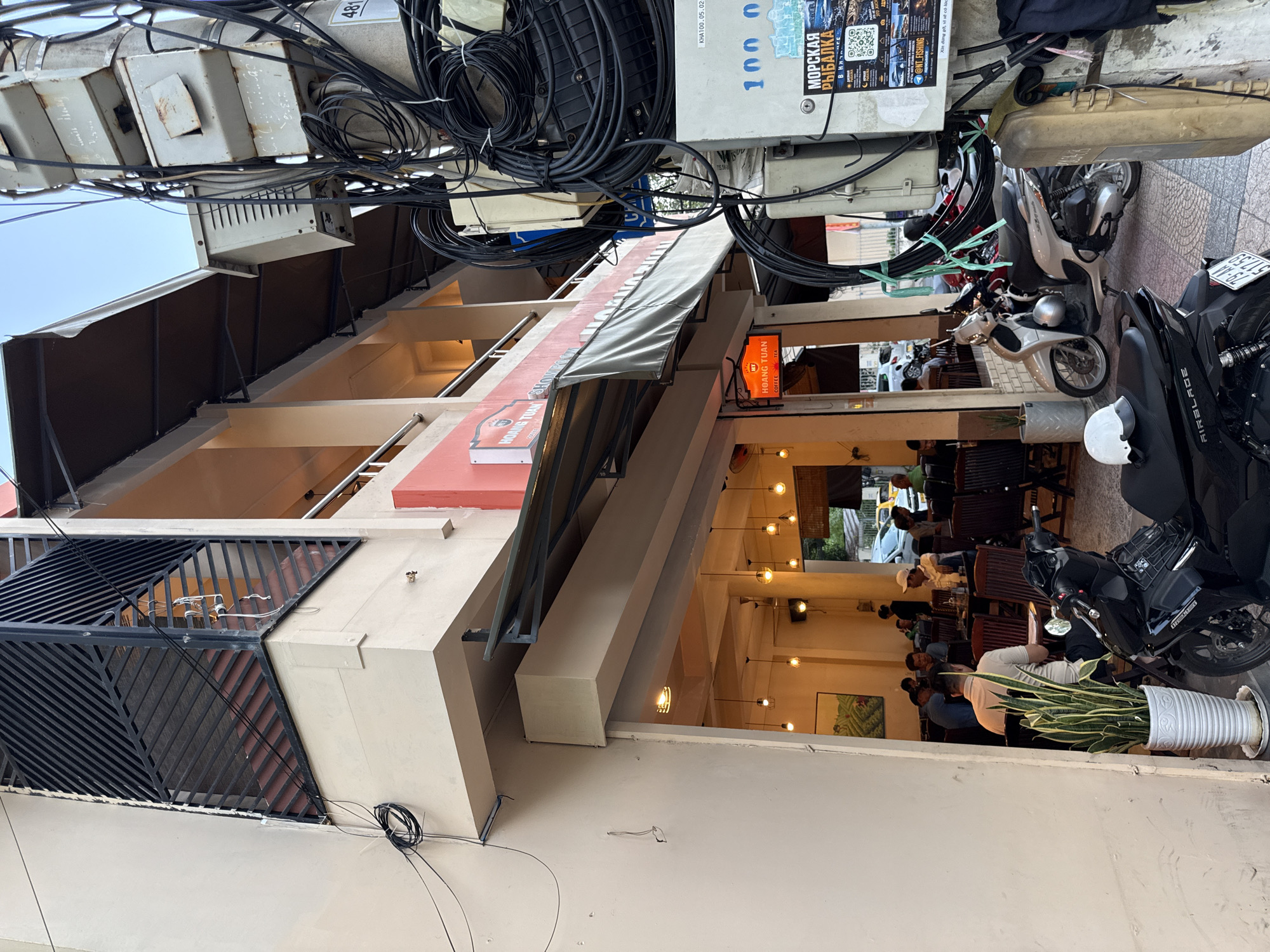 The image captures a street scene in Nha Trang, Vietnam, focusing on the exterior of a building with a coffee shop or restaurant. In the foreground, a black motorcycle is parked. The building itself has multiple levels, with balconies and awnings visible. A sign reading "HOANG TUAN COFFEE TEA" is prominent on the facade.
Inside the establishment, patrons are seated at tables, suggesting a busy atmosphere. The interior lighting, with exposed bulbs, creates a warm ambiance.
To the right of the building, a utility pole is heavily laden with electrical and telecommunication cables. Various electrical boxes and conduits are attached to the pole. A poster is affixed to one of the boxes, displaying Cyrillic text and images, possibly advertising a fishing trip.
The overall impression is a typical urban streetscape in Vietnam, blending commercial activity with the utilitarian infrastructure of the city.