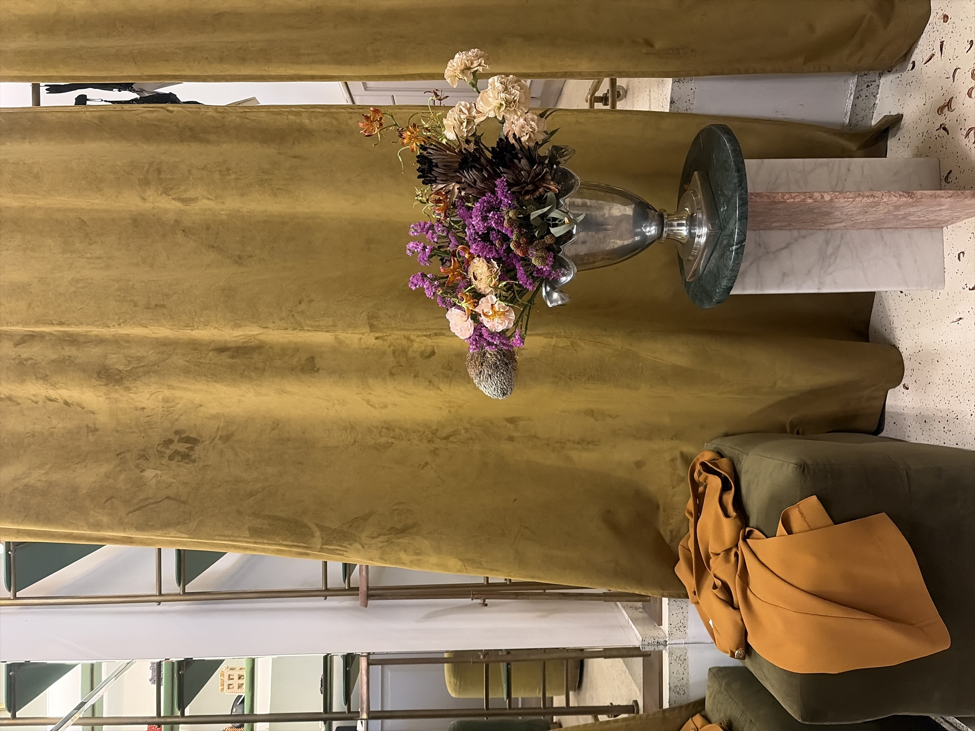 The image shows a still life arrangement within an indoor setting, likely a retail space or a well-decorated home. A metallic vase, featuring an ornate, fluted opening, is filled with a lush bouquet of flowers. The arrangement includes various types of flowers, with prominent purple statice, creamy white carnations, and some delicate orange and pink blossoms. Dark, spiky foliage adds texture and depth to the bouquet. Below the vase, a unique pedestal serves as a display. It's constructed from stacked blocks of different marble types, including a white marble base, a pinkish marble layer, and topped with a dark green marble circular plate. The pedestal itself sits on a speckled terrazzo floor. Behind the arrangement, a floor-to-ceiling curtain of lustrous, golden-ochre fabric drapes, creating a rich, warm backdrop. To the right of the pedestal, a portion of a dark green upholstered furniture piece is visible, with a crumpled, burnt-orange fabric draped over it. The overall impression is one of curated elegance and sophisticated interior design. The lighting appears to be artificial, casting soft shadows and highlighting the textures of the fabrics and flowers.