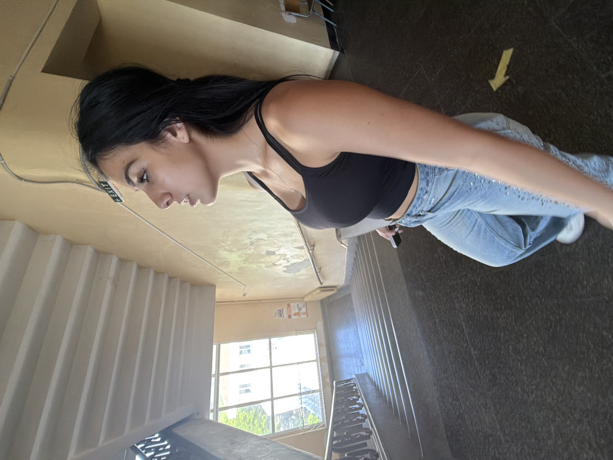 The image shows a young woman standing on a landing in an interior stairwell, likely within an older building in Burgas, Bulgaria. She has long, dark hair and is wearing a black, fitted crop top or tank top, and light blue jeans that appear to have some embellishments. A delicate silver necklace adorns her neck. She is captured in a posed moment, looking to her left and slightly downwards, holding a smartphone in her left hand.
The setting is a well-lit space, illuminated by natural light entering through a large, multi-paned window on the opposite side of the stairwell. Through this window, an exterior building with multiple windows and some greenery is visible, indicating it is daytime. The walls are a light beige or tan, displaying some visible wear. A partially obscured "EXIT" sign, with white text on a dark background, can be seen on the upper left wall. The floor is dark gray or brown with a textured pattern, and a bright yellow arrow pointing downwards is painted on it, likely for directional guidance. The stairwell features several flights of stairs with white or light-colored risers and dark treads, and a banister is visible. The ceiling is simple, with some conduits or pipes running across it, contributing to the older building aesthetic.