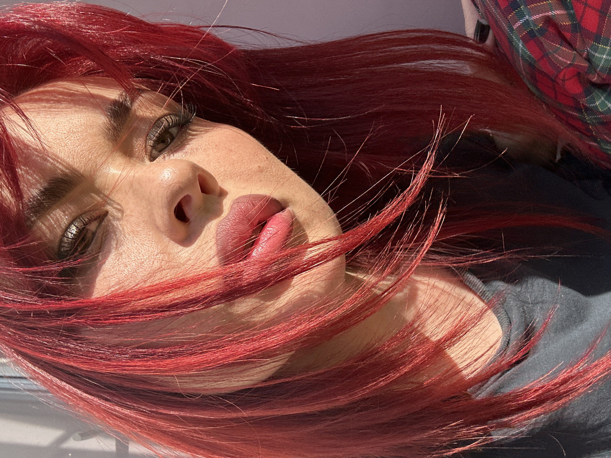 The image shows a close-up, chest-up portrait of a young woman with bright red hair, illuminated by sunlight casting shadows across her face. She is looking towards the camera with her head tilted. Her makeup includes dark eyeliner and mascara, and her lips are painted with a glossy, pink-toned lipstick. She is wearing a dark gray t-shirt and partially visible is a plaid garment with red, green, and white checks. The background suggests an indoor environment with a window or blinds on the left, and a plain wall or surface on the right. There is no discernible connection to the city of Узюково, Russia, within the visual information provided in the image.