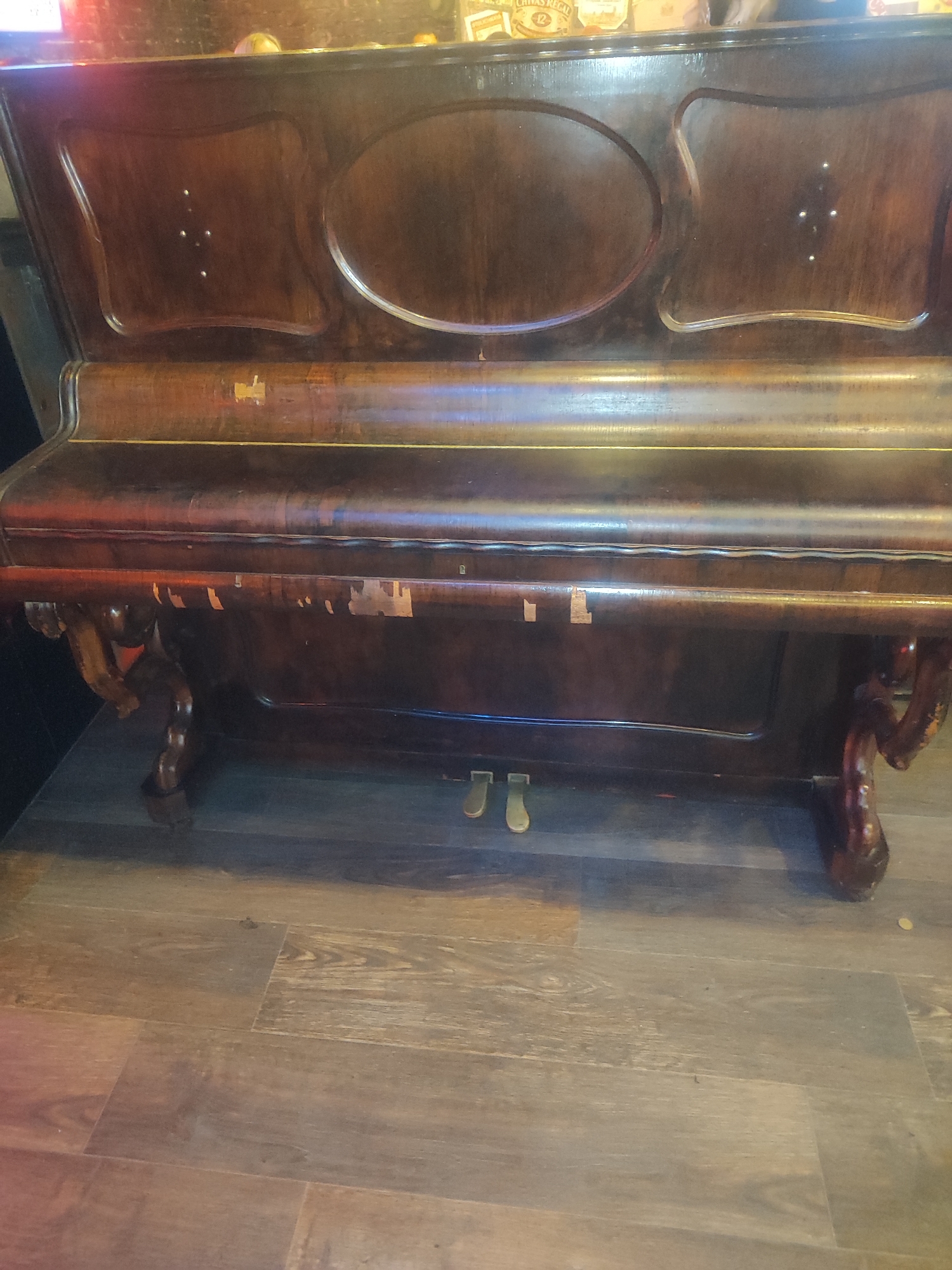 An upright piano is shown in a well-lit interior. The piano is made of dark wood and has ornate carvings on its legs. The surface of the piano shows signs of wear and tear, with some areas of chipped paint. There are two pedals visible on the floor in front of the piano. The piano is set against a wooden floor. The background is blurred, but suggests a dimly lit environment, possibly a bar or restaurant.
