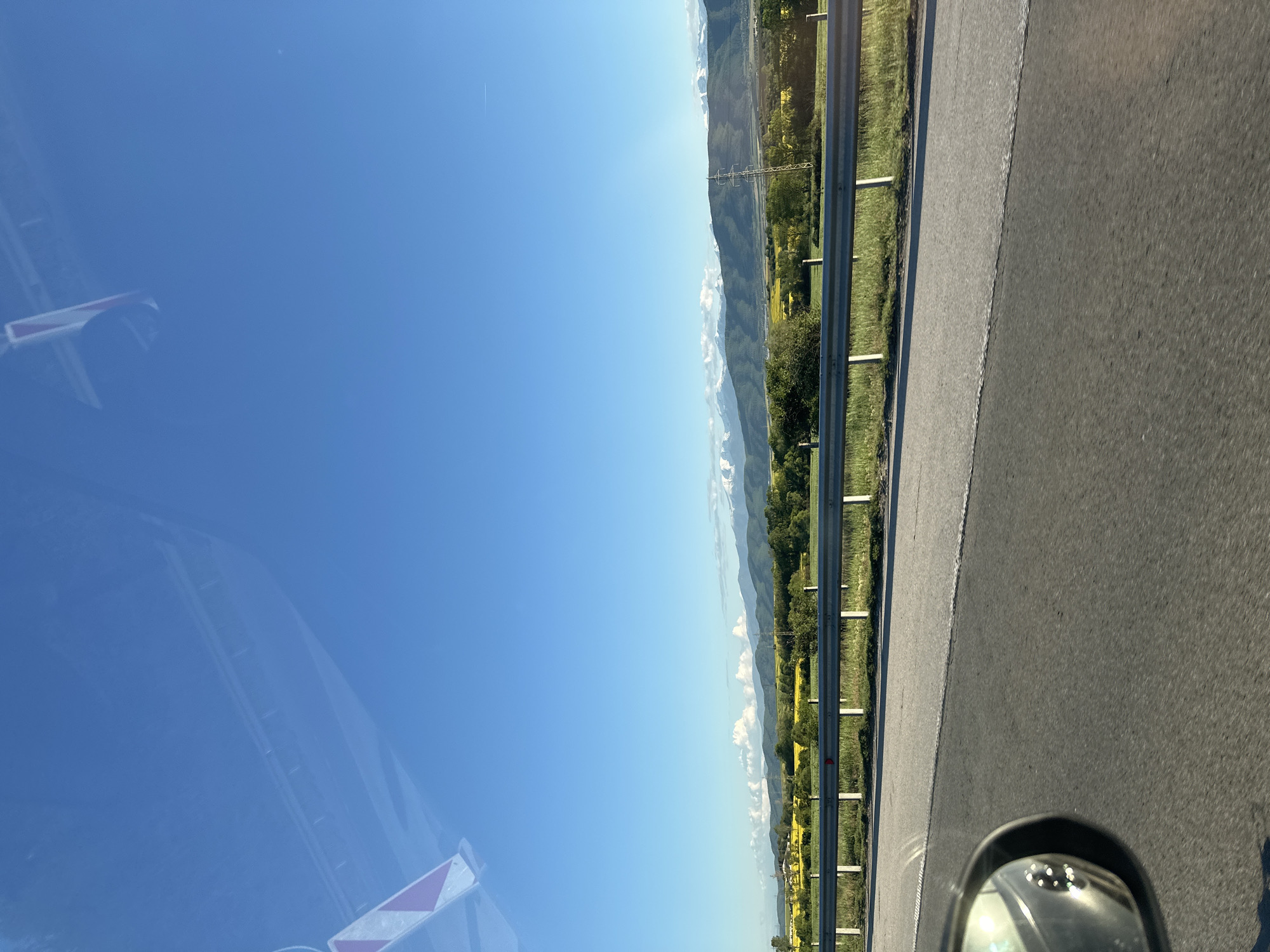 The image depicts a view from a moving vehicle on a highway, likely in a mountainous region near Rajana, Bulgaria. The sky is clear blue, occupying a significant portion of the frame. To the right, a guardrail runs along the side of the road. Beyond the guardrail is a landscape with trees, grassy areas, and mountains in the distance. The asphalt road takes up a portion of the lower right corner, and a side-view mirror of the vehicle is visible in the foreground. In the left corner of the shot, a traffic sign, blurred by the car's window, is partially visible.