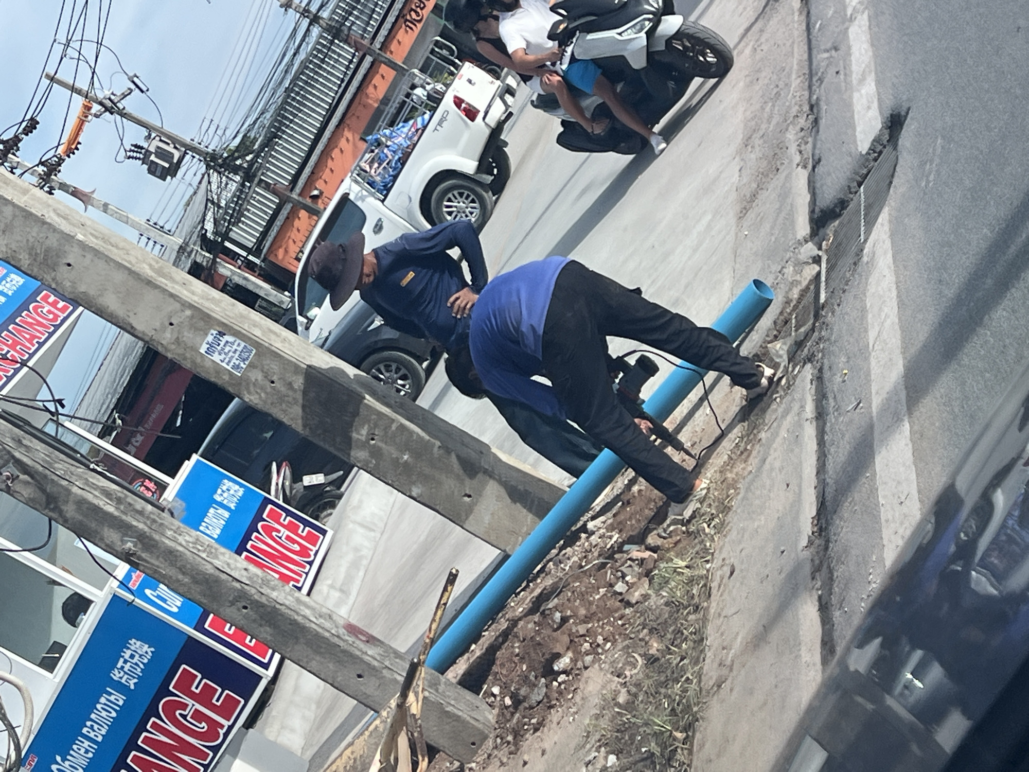 Here is a description of the image:
Two men are shown near the side of a road in Baan Bang Rak, Thailand. One man is wearing a hat and a dark blue shirt. The other man is wearing a light blue shirt. They appear to be working together on something near a blue pipe that runs alongside the sidewalk. A white pickup truck and a motorcycle with two people are parked nearby. Utility poles and overhead wires are also visible. There are "EXCHANGE" signs with text in multiple languages near the poles. Part of a car is visible in the foreground, suggesting the image was taken from inside a vehicle.