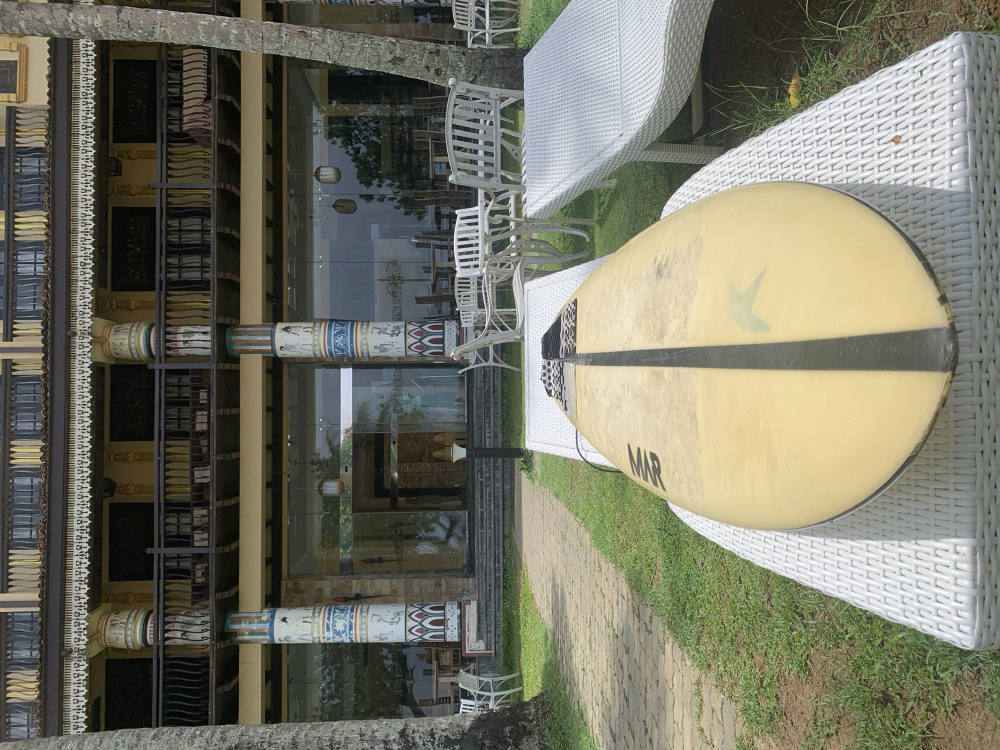 The image shows a surfboard resting on white wicker furniture on a lawn. In the background, a building with decorative columns is visible. The location is Kathaluwa, Sri Lanka.