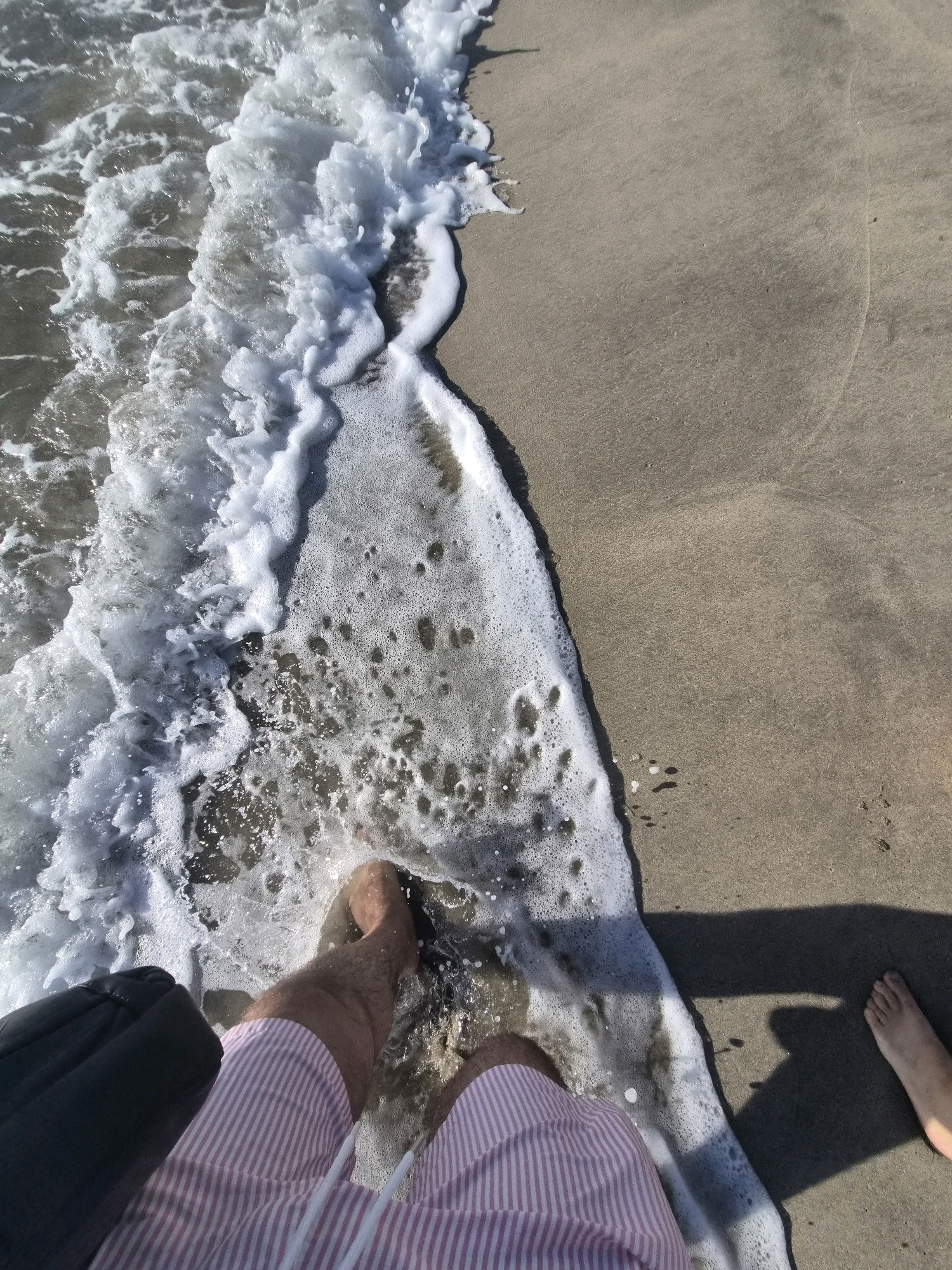 A first-person perspective shot captures a wave washing over sand at Esposende, Portugal. The foreground shows a person's legs from the knees down, clad in pink and white striped shorts. To the left of the legs is a dark, rectangular object, possibly a bag. The person's feet are submerged in the foamy water. Another person's foot is visible in the lower right corner of the frame, casting a shadow on the sand. The environment is a sandy beach with the ocean's edge.