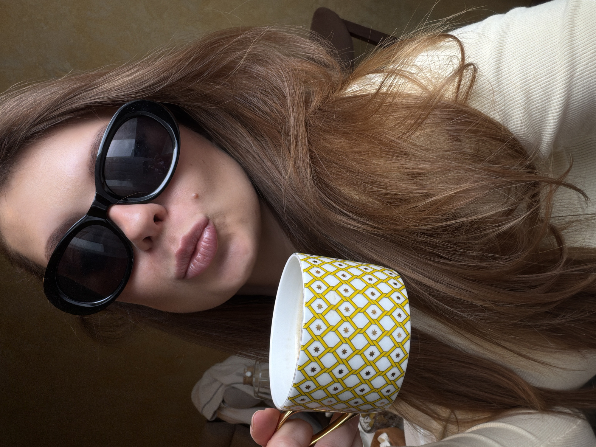 The image shows a woman with long brown hair, wearing large black sunglasses, and holding a white mug with a yellow and white geometric pattern. The mug appears to contain coffee. She is wearing a cream-colored top. The background is a dark, out-of-focus surface. There is no information in the image indicating this was taken in Khimki, Russia.