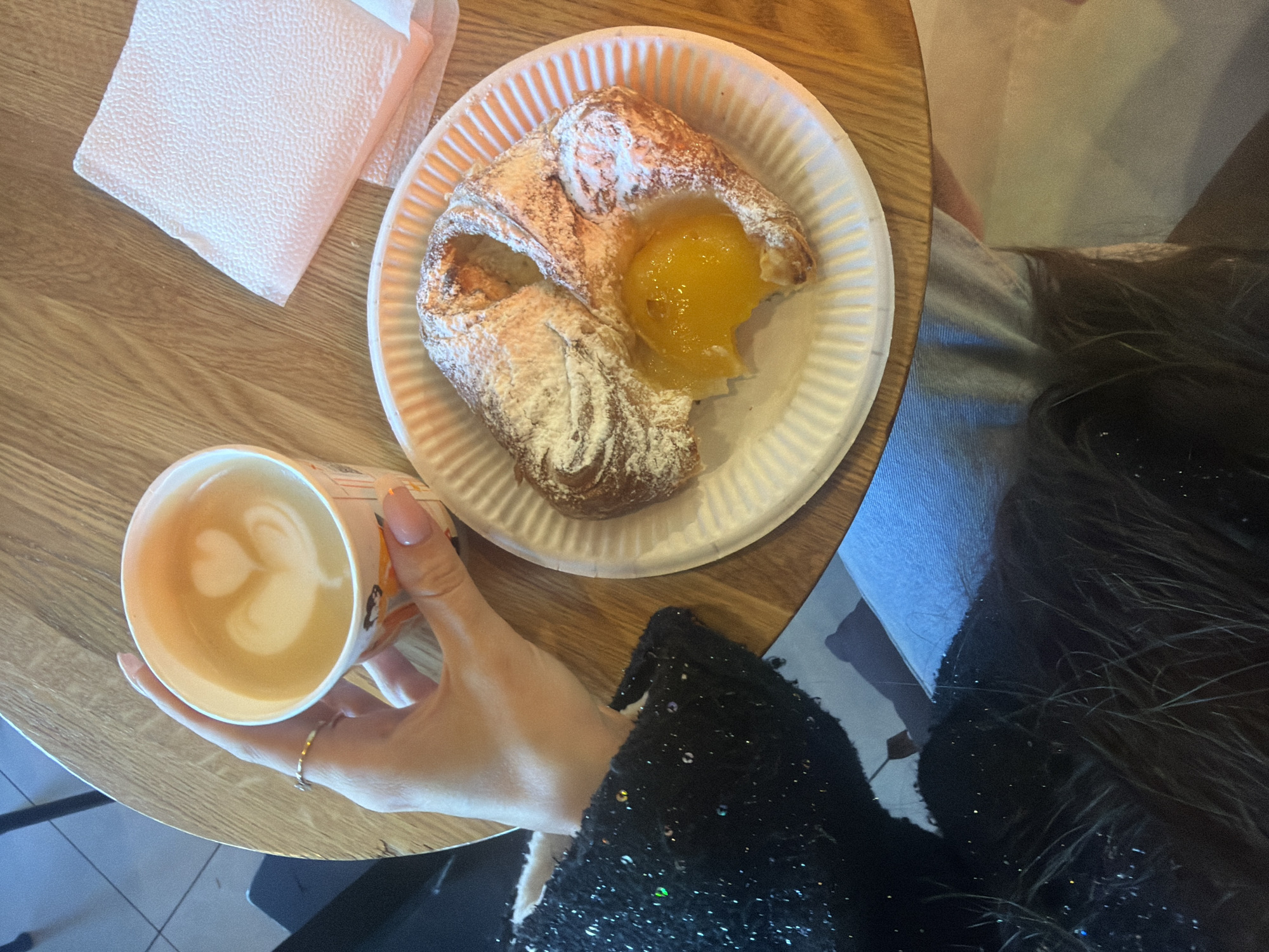 The image shows a table at an establishment, possibly a cafe or bakery, in Saint Petersburg, Russia. A person's hand is holding a cup of latte with a heart-shaped foam design. Next to the cup, there is a pastry filled with yellow jam on a white paper plate. A stack of white napkins is also present on the table. The person is wearing a black, sequined jacket. The table has a light wood grain finish. Part of a blue garment (jeans) and a dark coat are visible in the background, suggesting the person is seated.