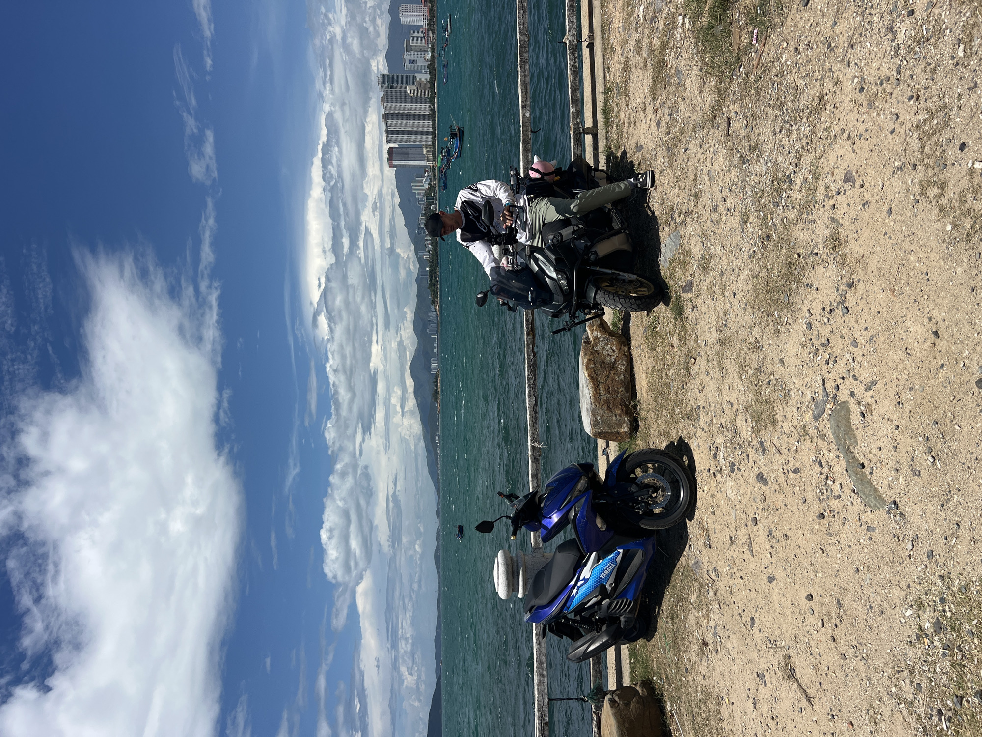 The image shows a coastal scene in Bac Nha Trang Ward, Vietnam. Two motorcycles are parked near a waterfront. A man is seated on the rear motorcycle. The background includes a body of water, a city skyline, and a partly cloudy sky. The ground surface appears to be a mix of dirt and gravel.