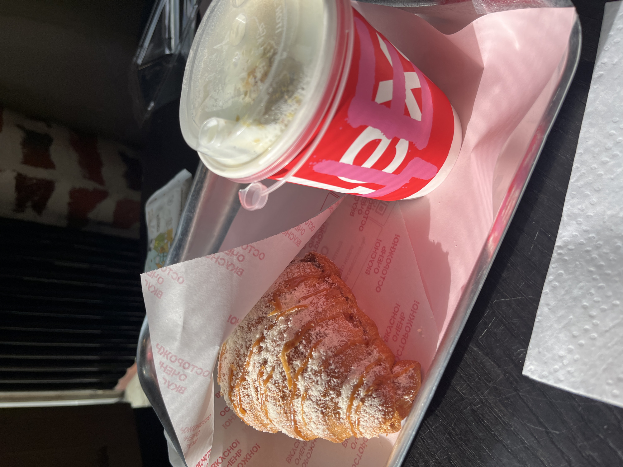The image shows a tray containing a red and white cup of liquid with a clear plastic lid, and a croissant dusted with powdered sugar and drizzled with caramel. They sit on a piece of pink paper with text in Russian. The tray is on a dark surface, possibly a table. The image may have been taken in Shushary, Russia given the text on the pink paper.